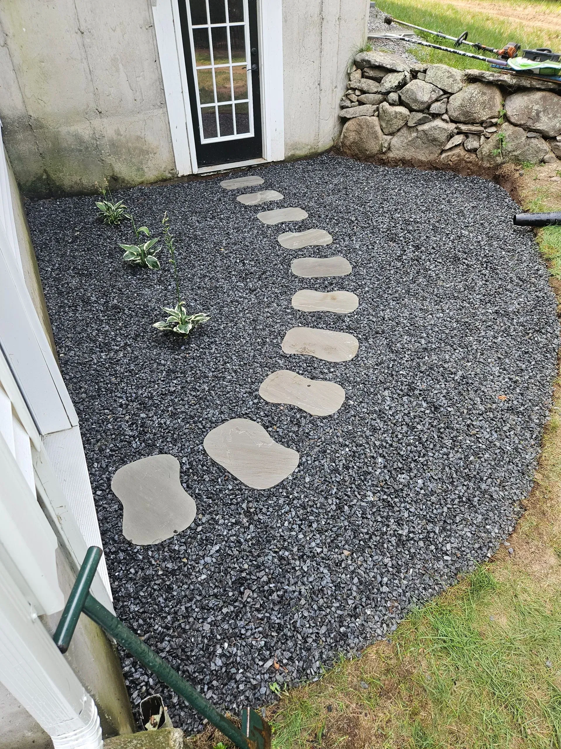 Curved gravel path with stepping stones leading to a black door beside a stone wall.