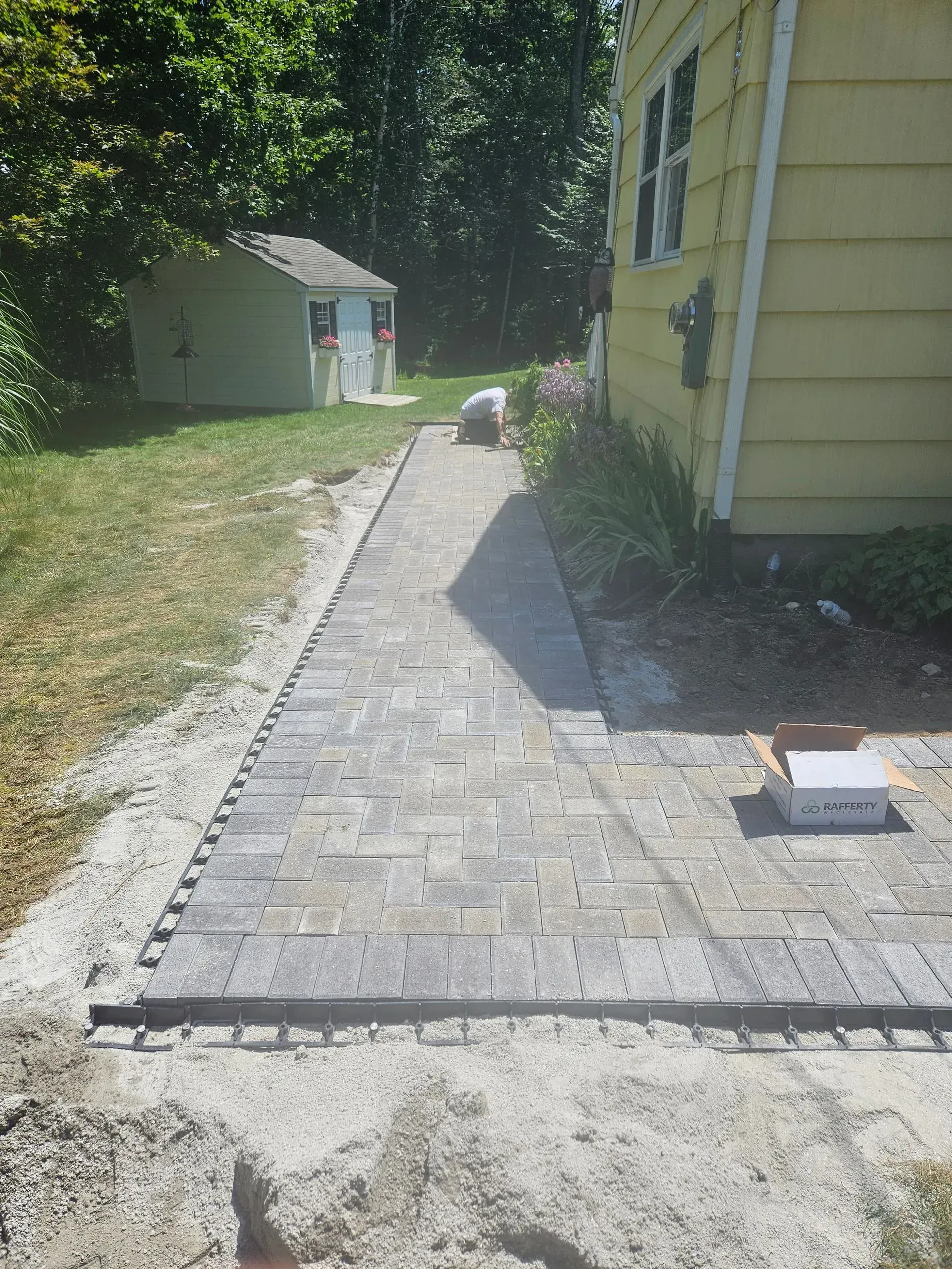 Brick walkway under construction beside a yellow house, with gravel and a shed in the yard.