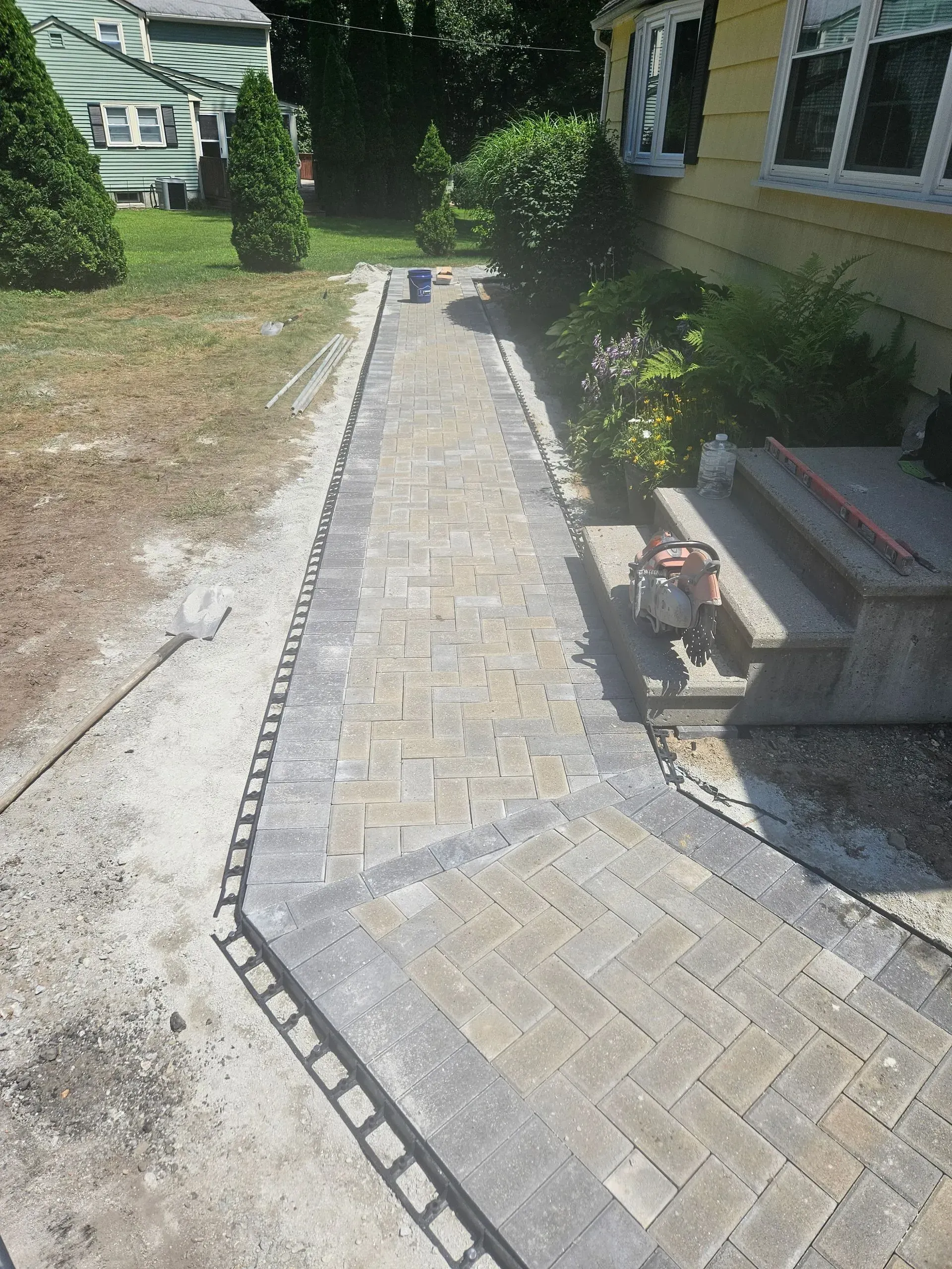 Brick walkway along a house with a small dog on the steps, bordered by grass and shrubs.