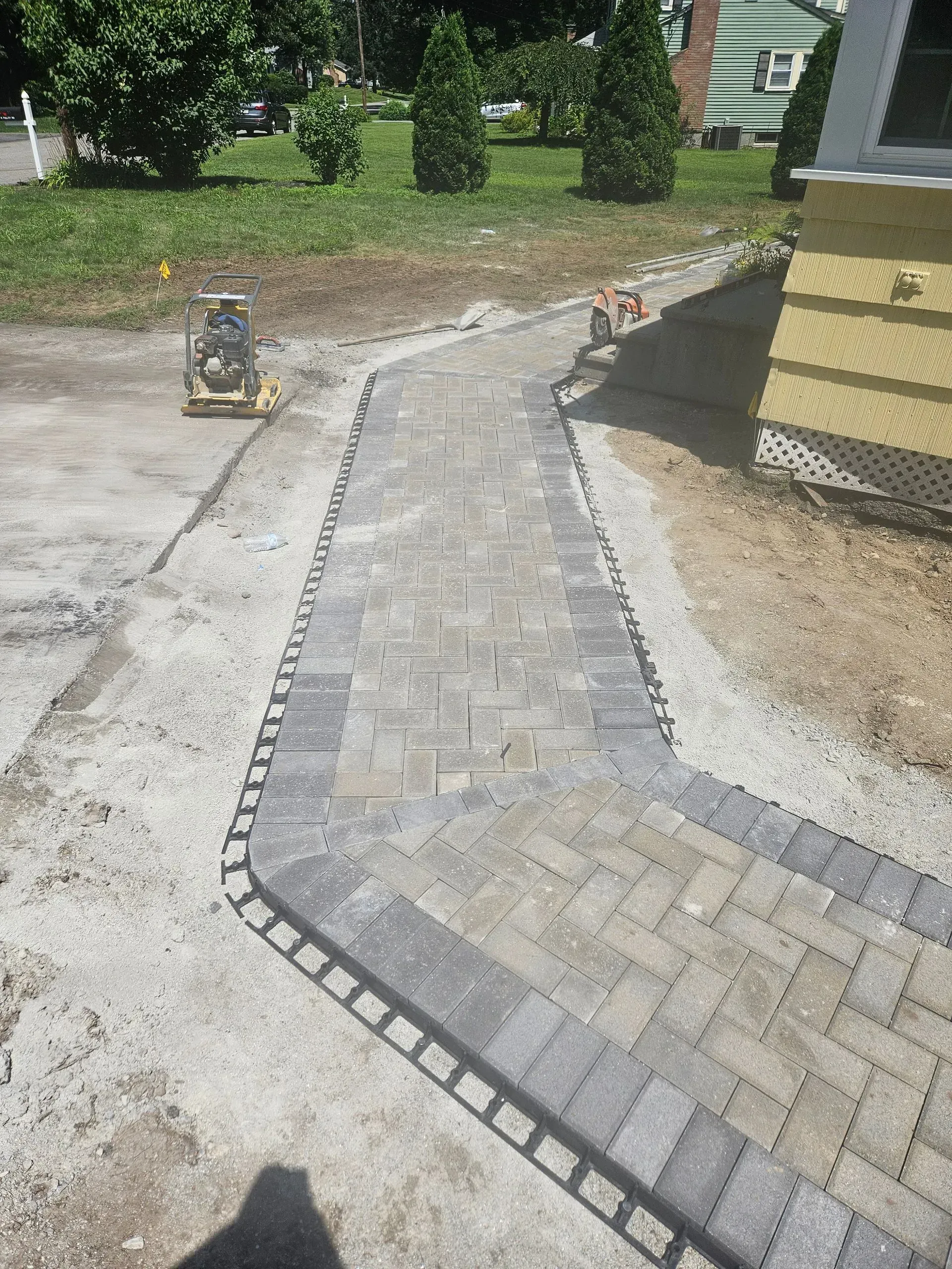 New gray paver walkway under construction beside a house, with gravel edges and a wheelbarrow nearby