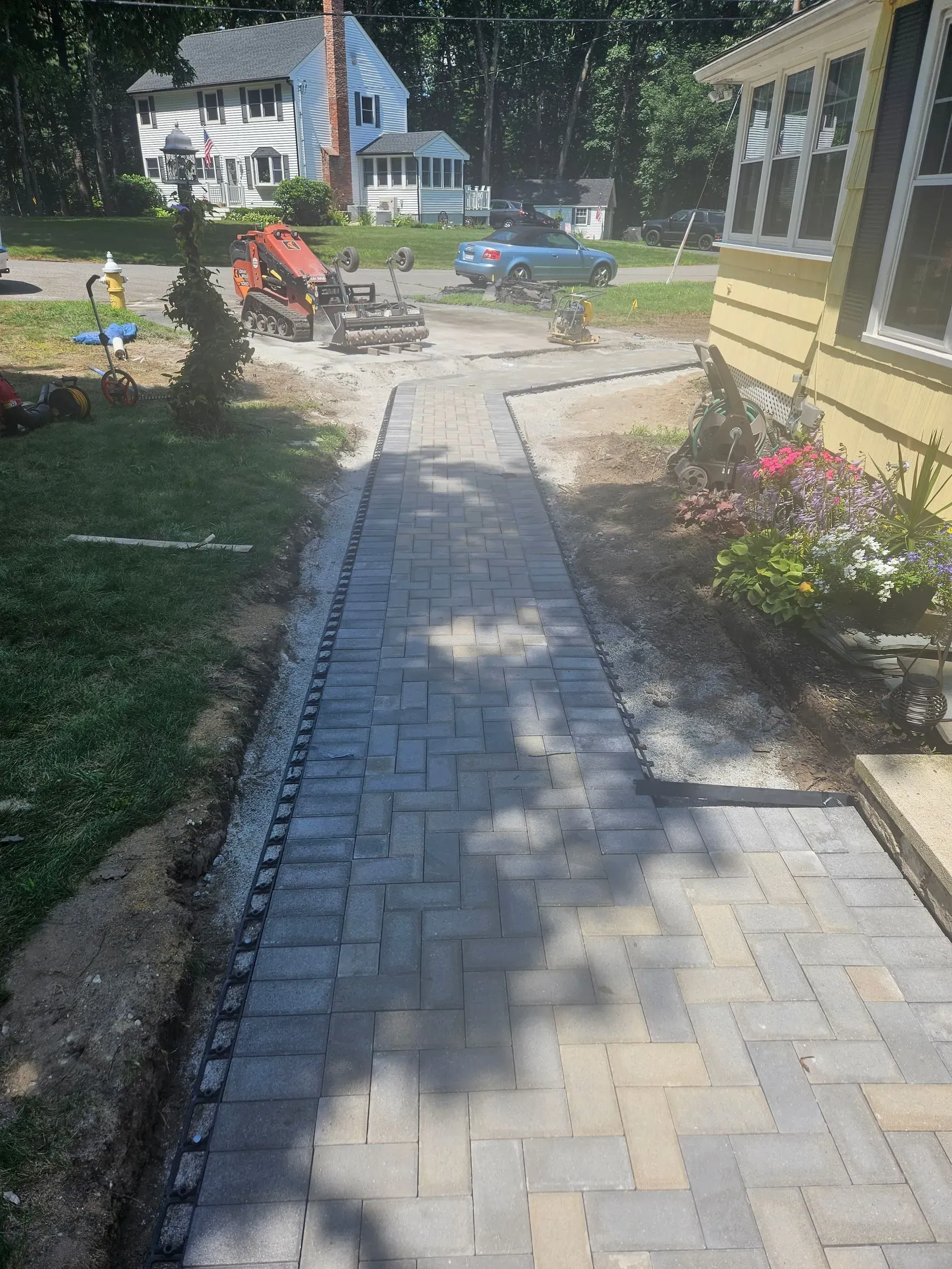 Stone walkway with pavers leading through a sunny garden beside a yellow house and flowers