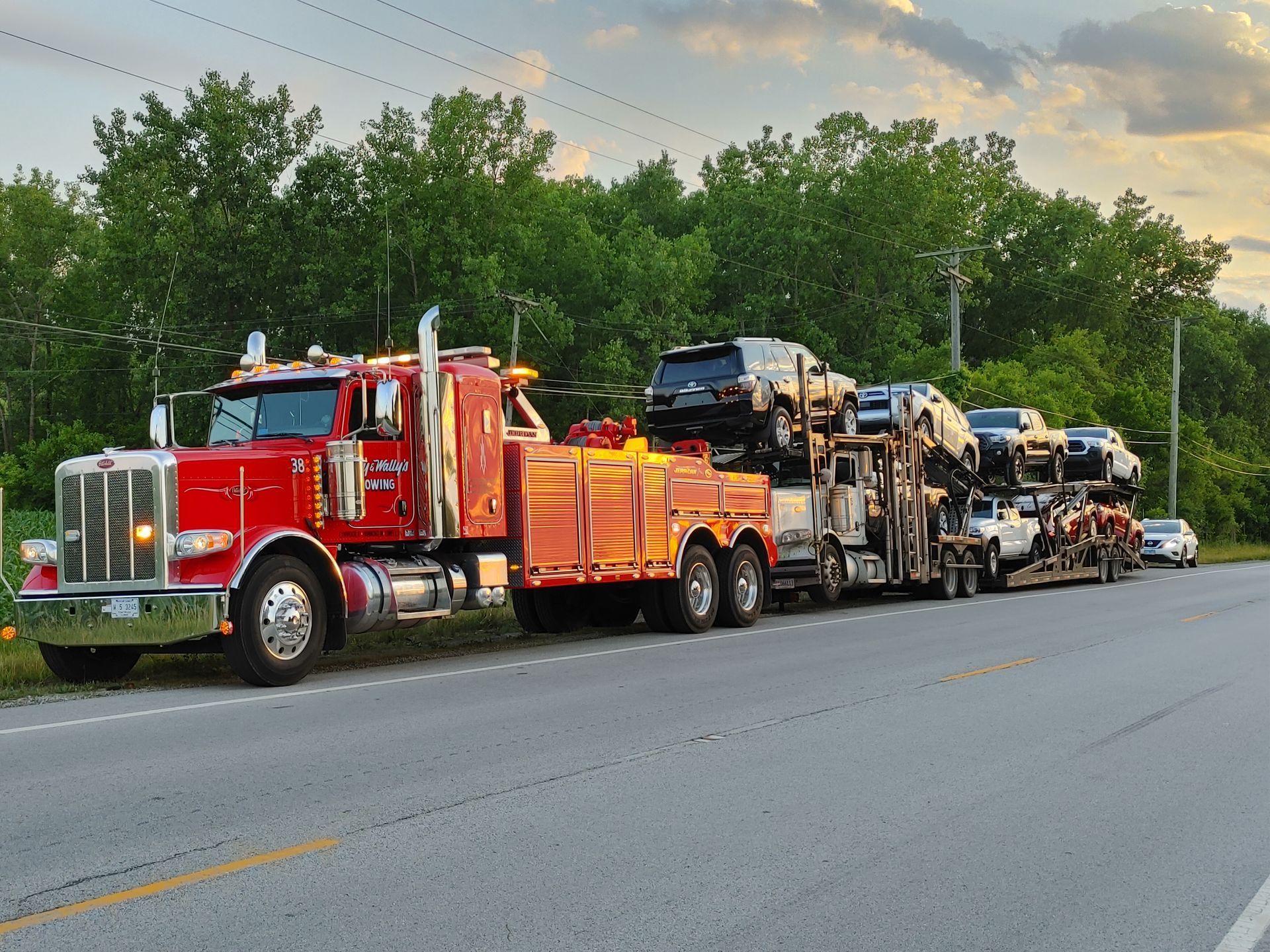A red tow truck carrying several SUVs on a flatbed trailer parked along a rural road at sunset.