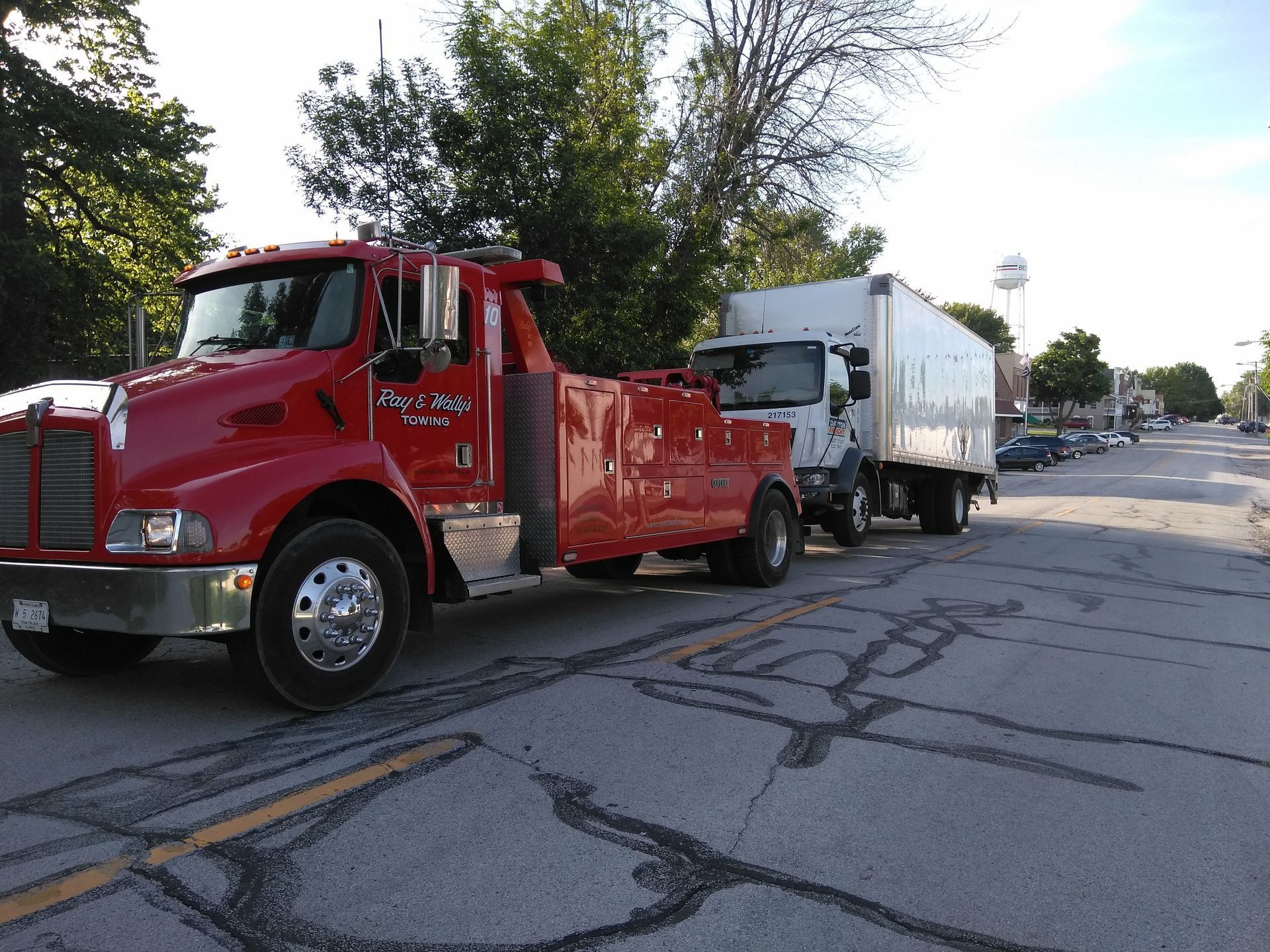 Red tow truck towing a white box truck on a street.