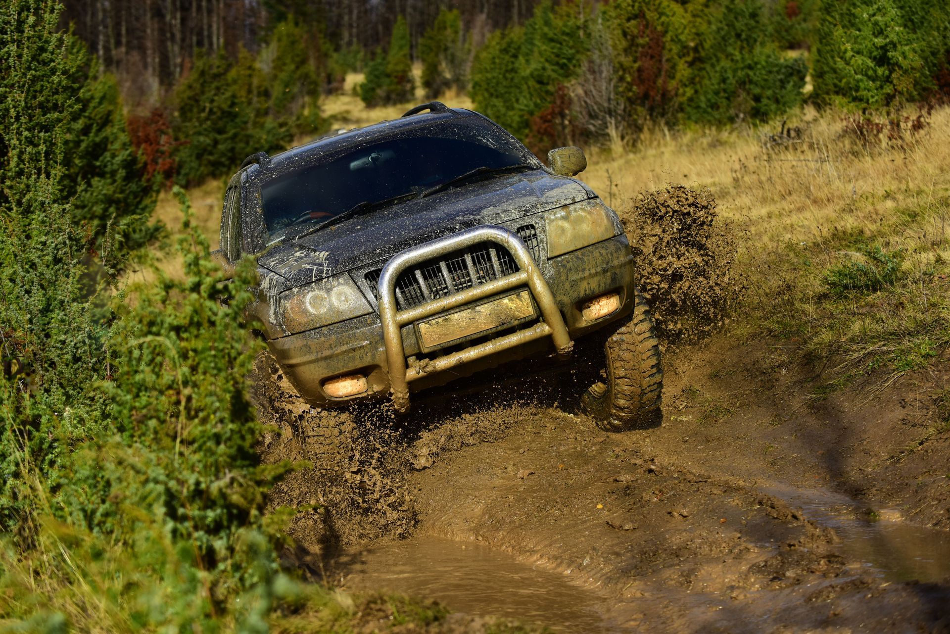 Dark SUV driving through mud on a dirt path in a wooded area, splashing muddy water.