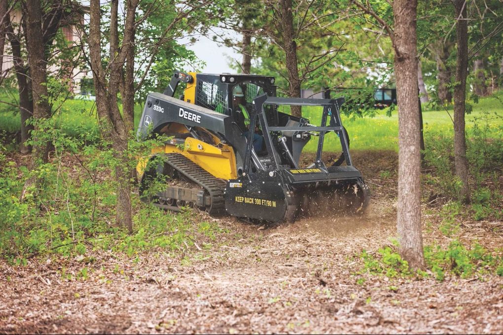 Black skid steer clearing brush in a wooded area, kicking up dirt among trees