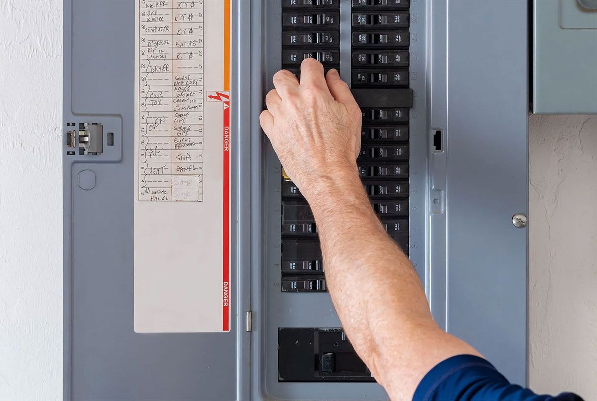 Hand reaching into an open electrical breaker panel with labeled circuit map inside