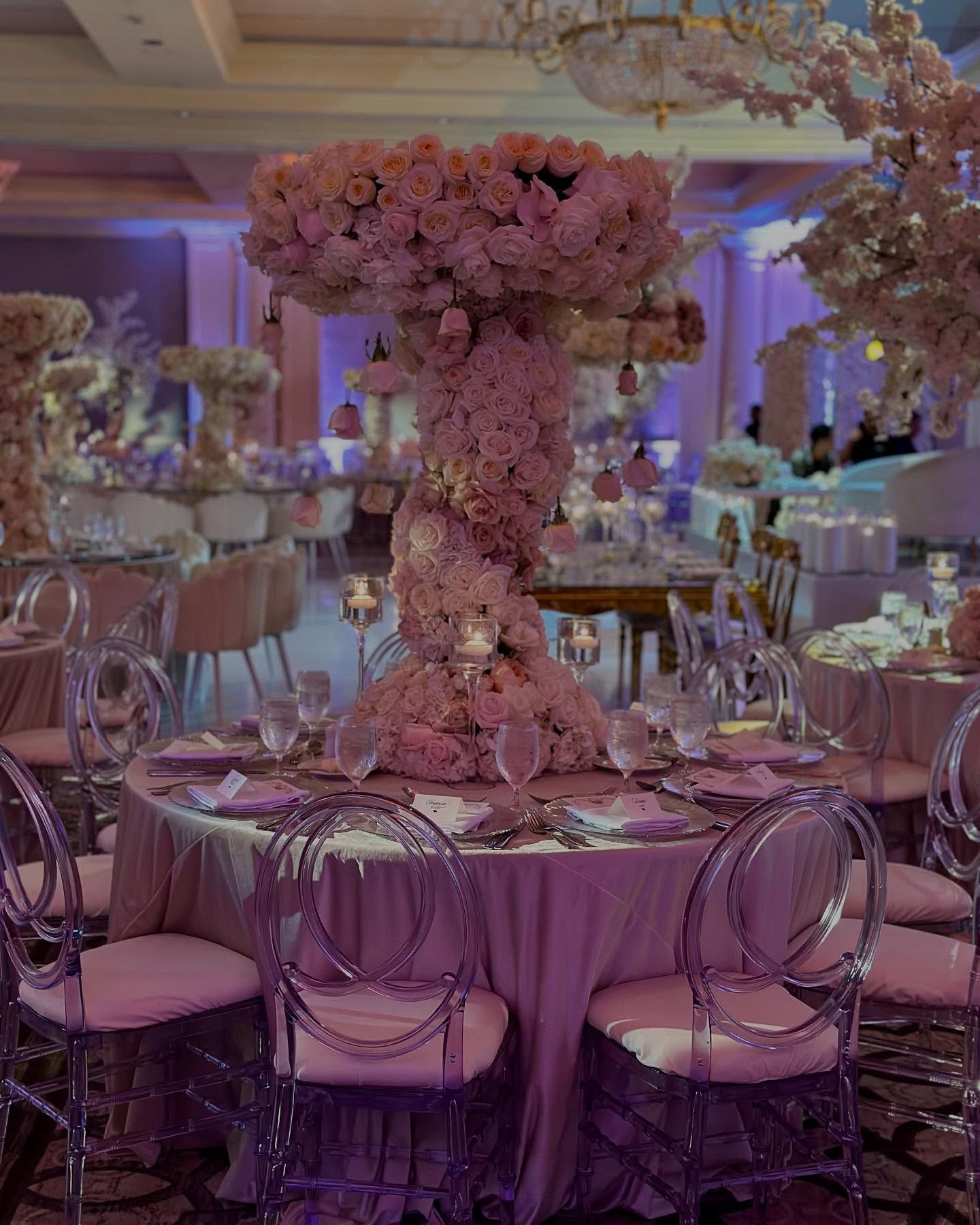 Elegant wedding reception with floral centerpiece, round tables, and clear chairs.