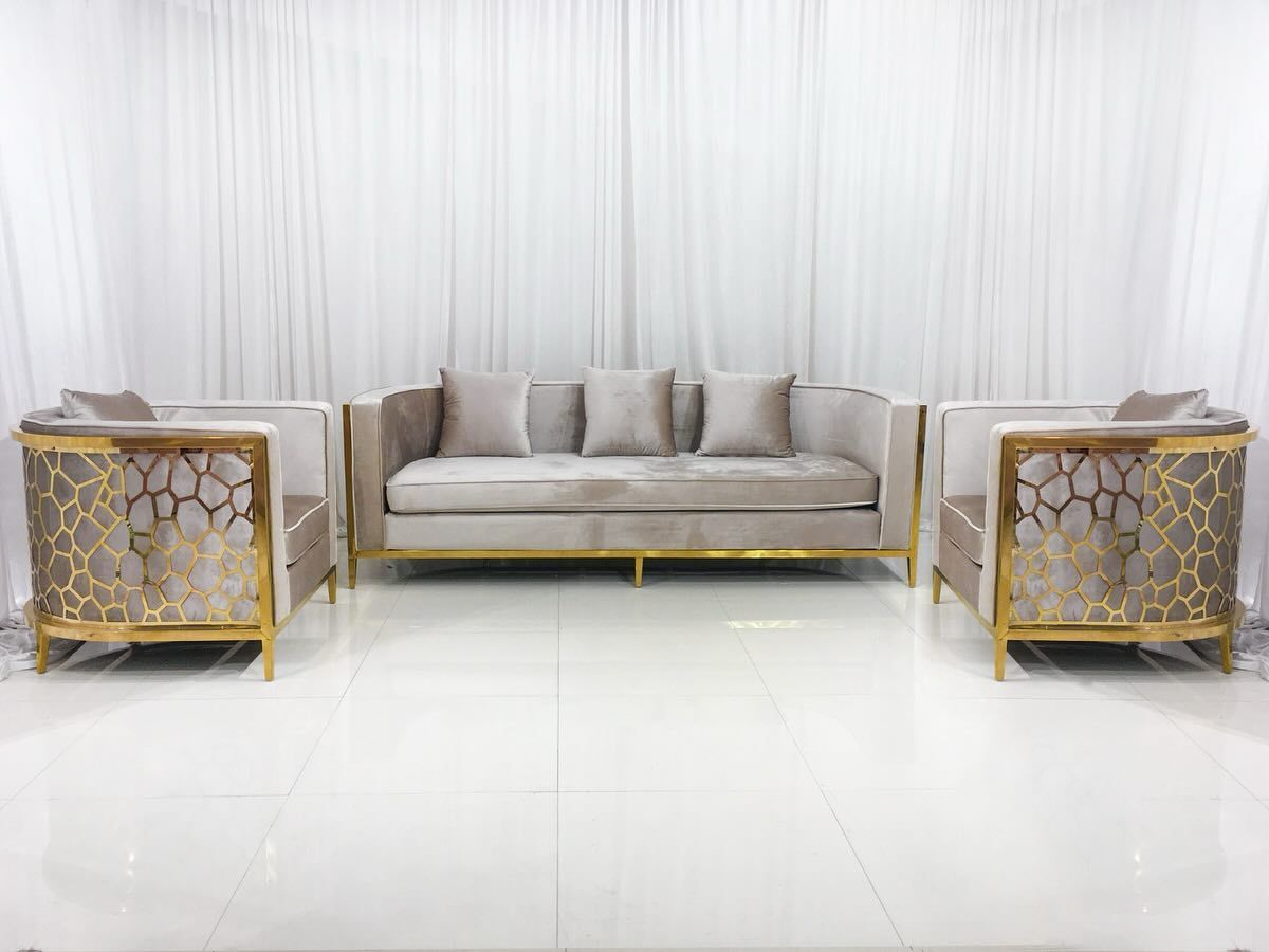 A modern living room set with a gray sofa and two armchairs, all with gold trim, set against white drapes.