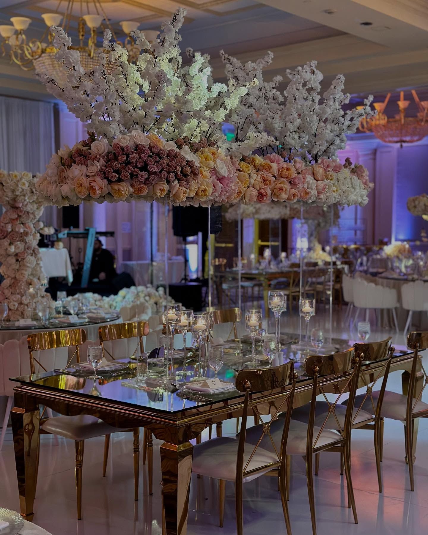 Formal dining room with a long mirrored table set for a meal, beneath large flower arrangements.