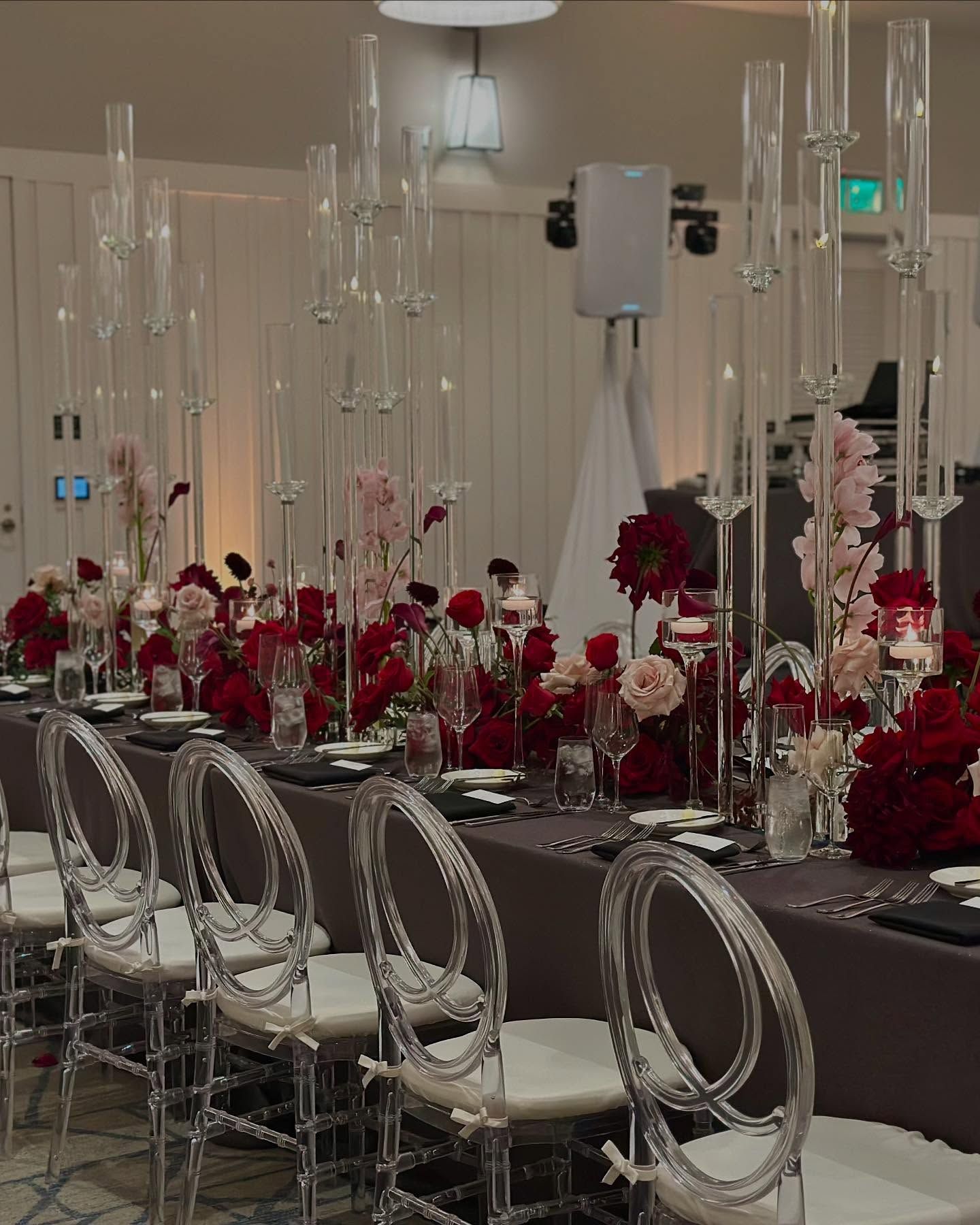 Long dining table set for a formal event with red floral arrangements, clear chairs, and tall candle holders.