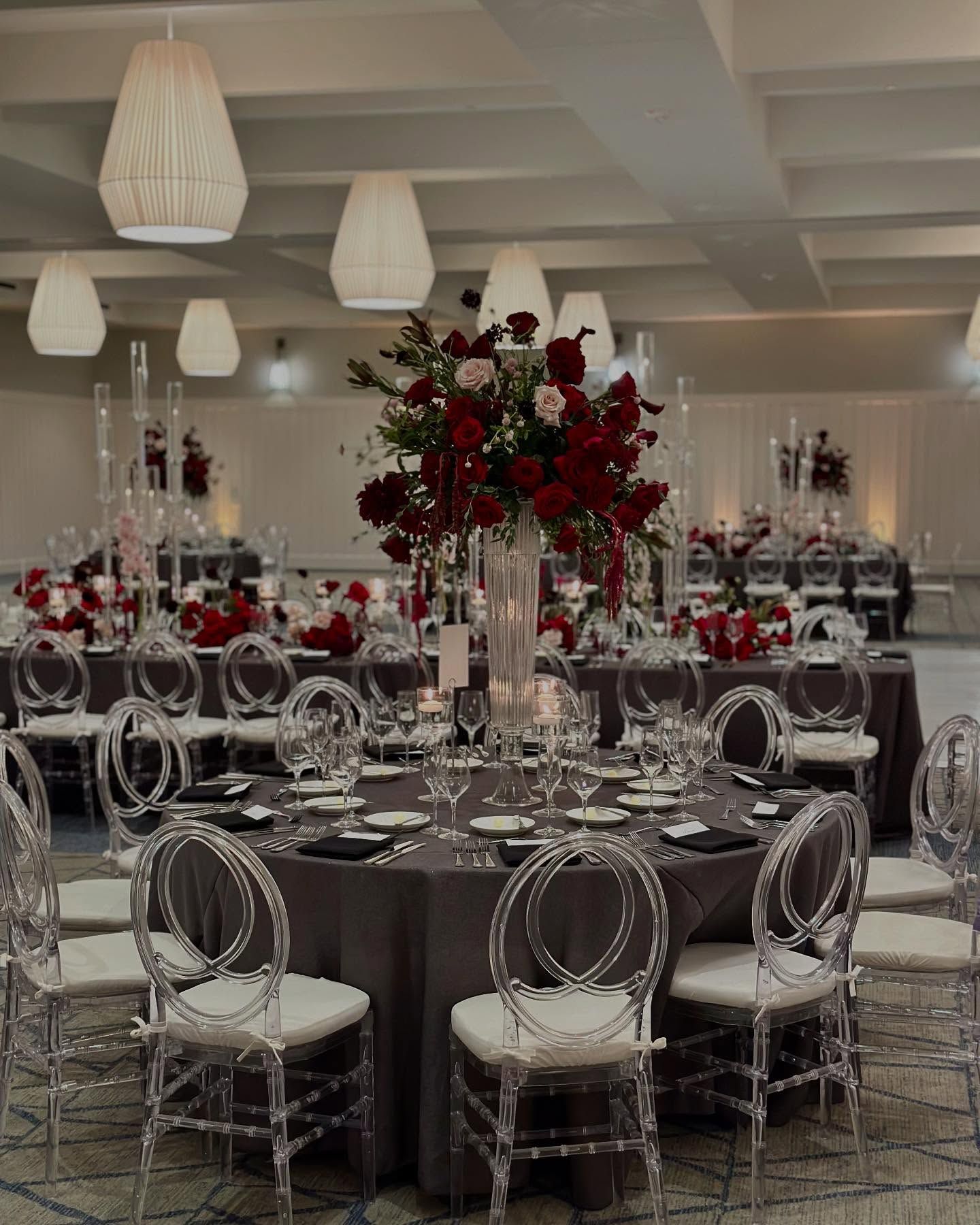 Elegant reception hall with round tables set for a formal event. Red floral centerpieces, clear chairs.
