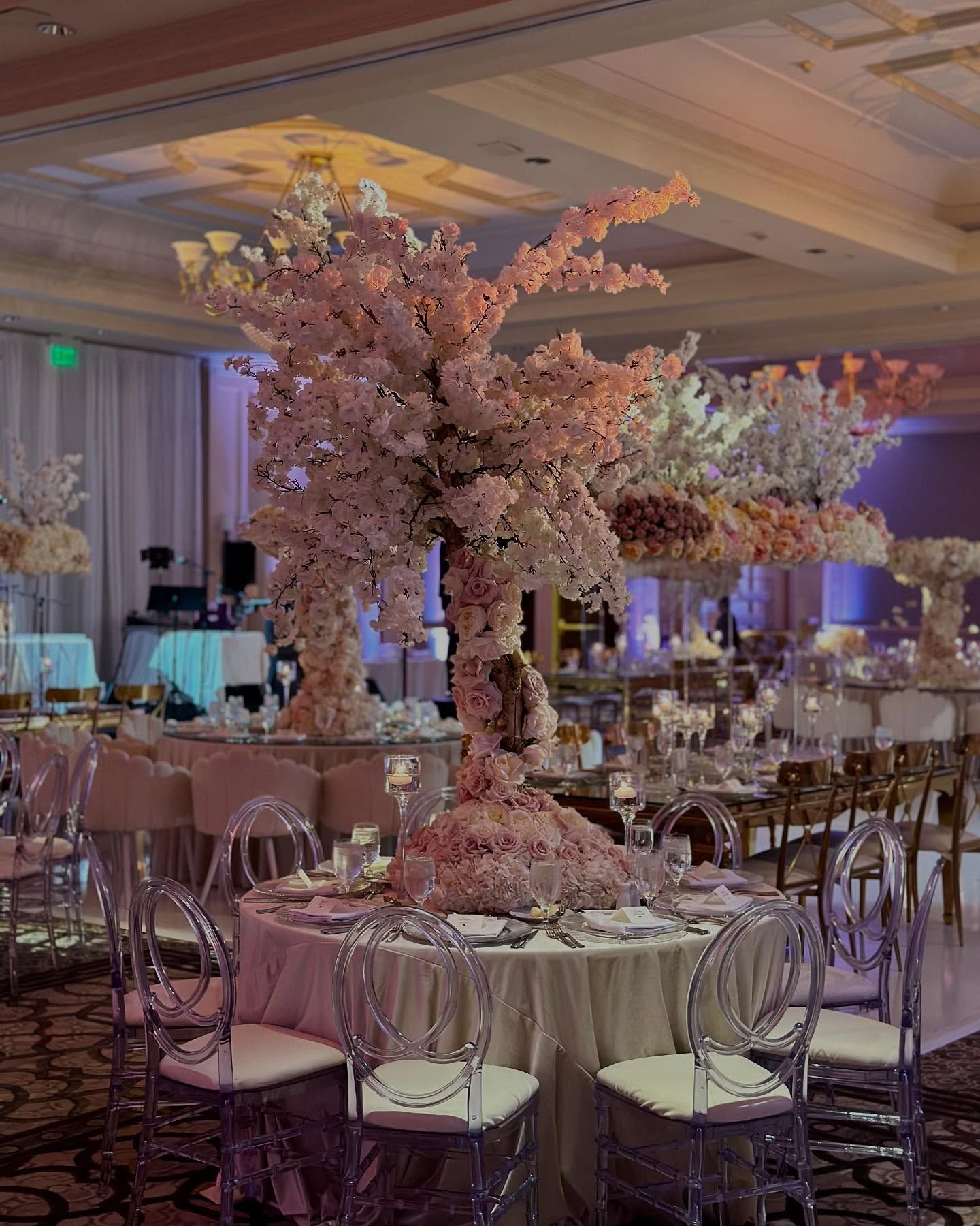 Elegant wedding reception with pink floral centerpieces and clear chairs around round tables.