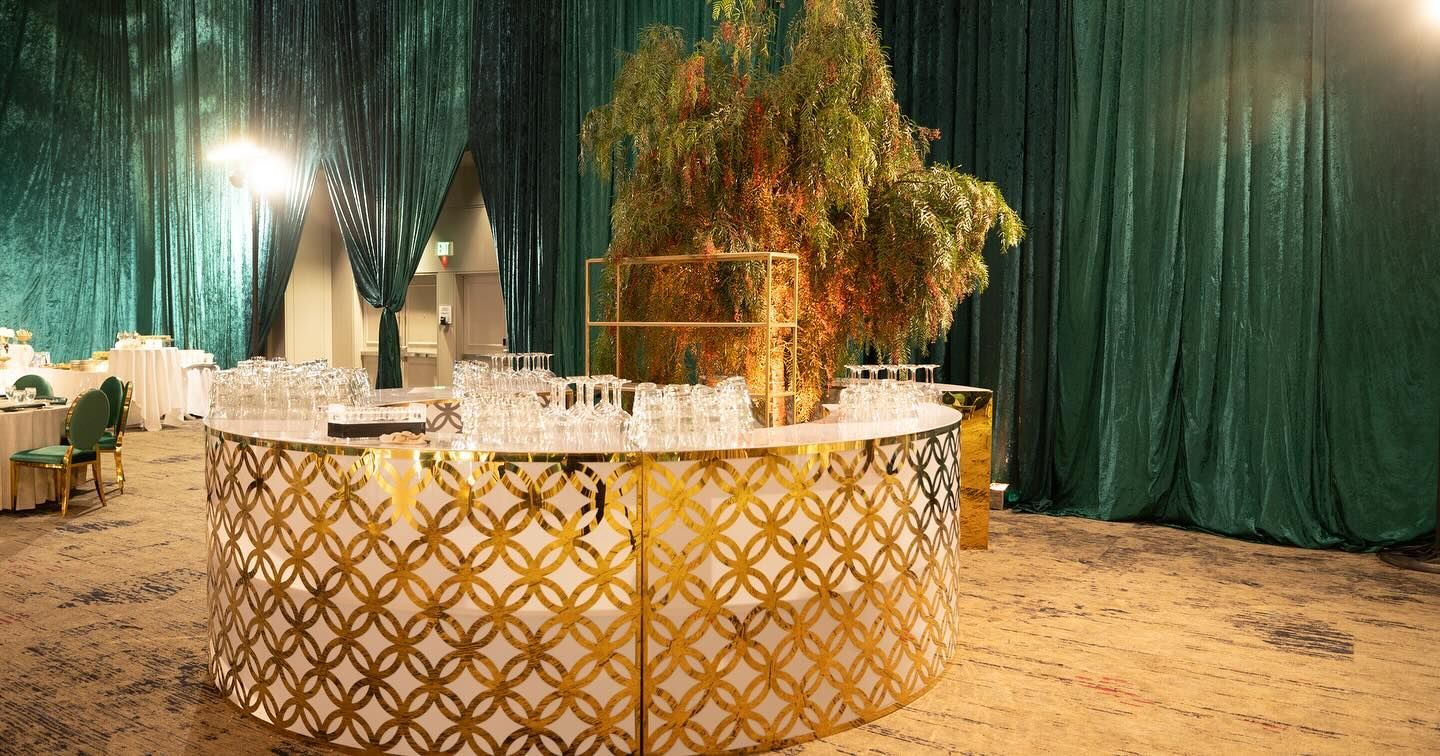 Golden patterned bar with glassware, green backdrop, and leafy tree in a formal setting.