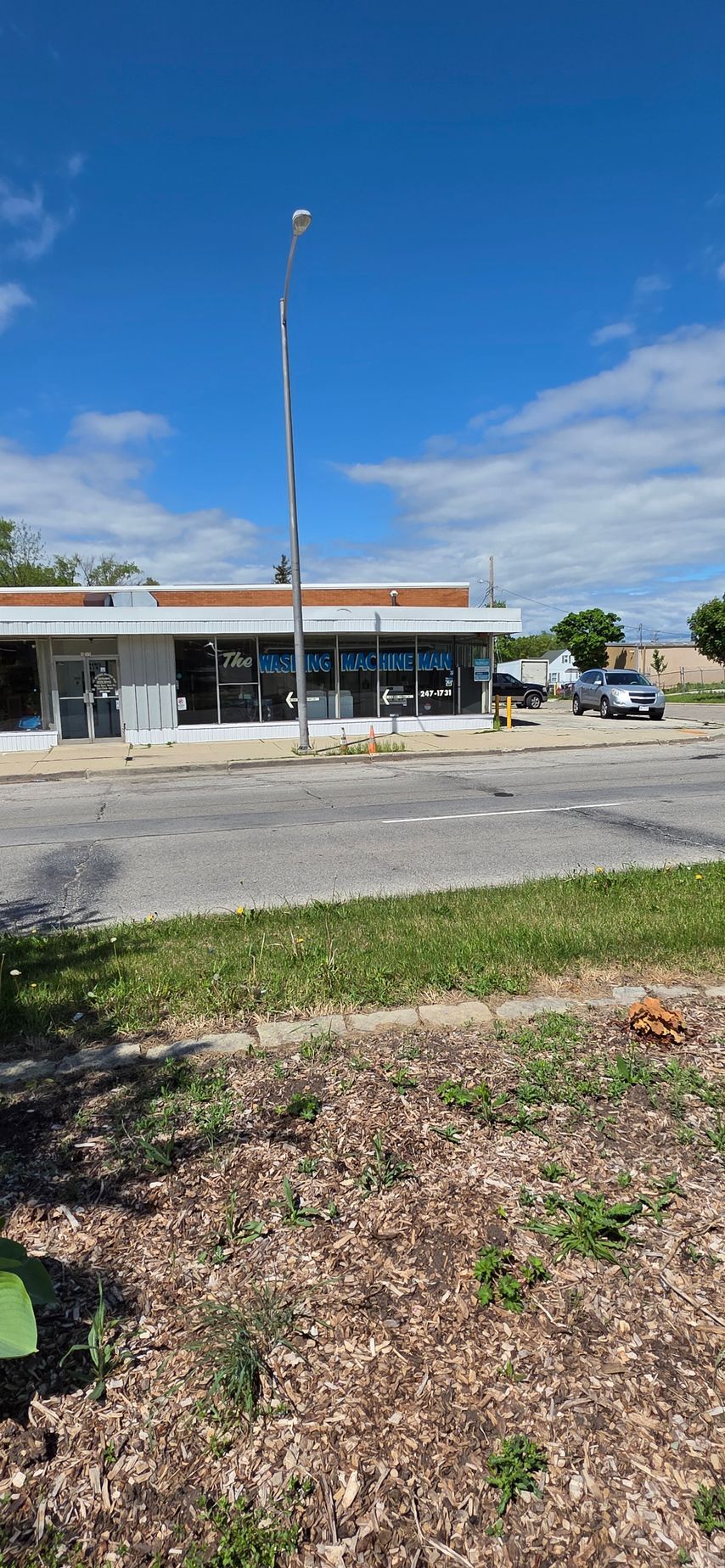 Low roadside storefront with blue awnings, tall flagpole, parked SUV, and grassy foreground under a blue sky