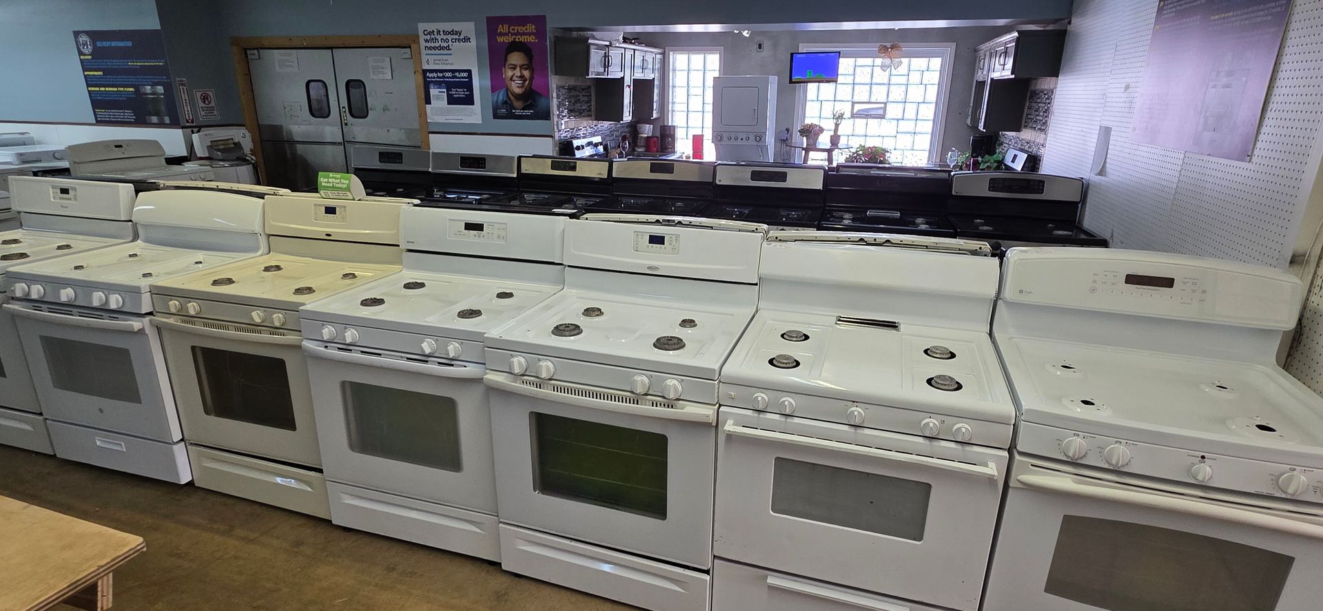 A row of white stoves and ovens in a showroom or store, with shelves and posters in the background.