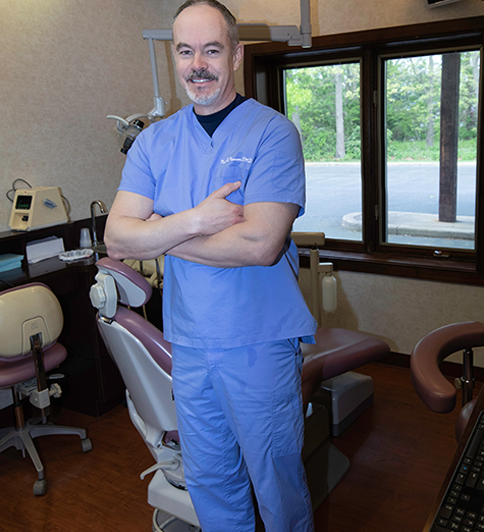 Dentist in blue scrubs standing with arms crossed in a dental office