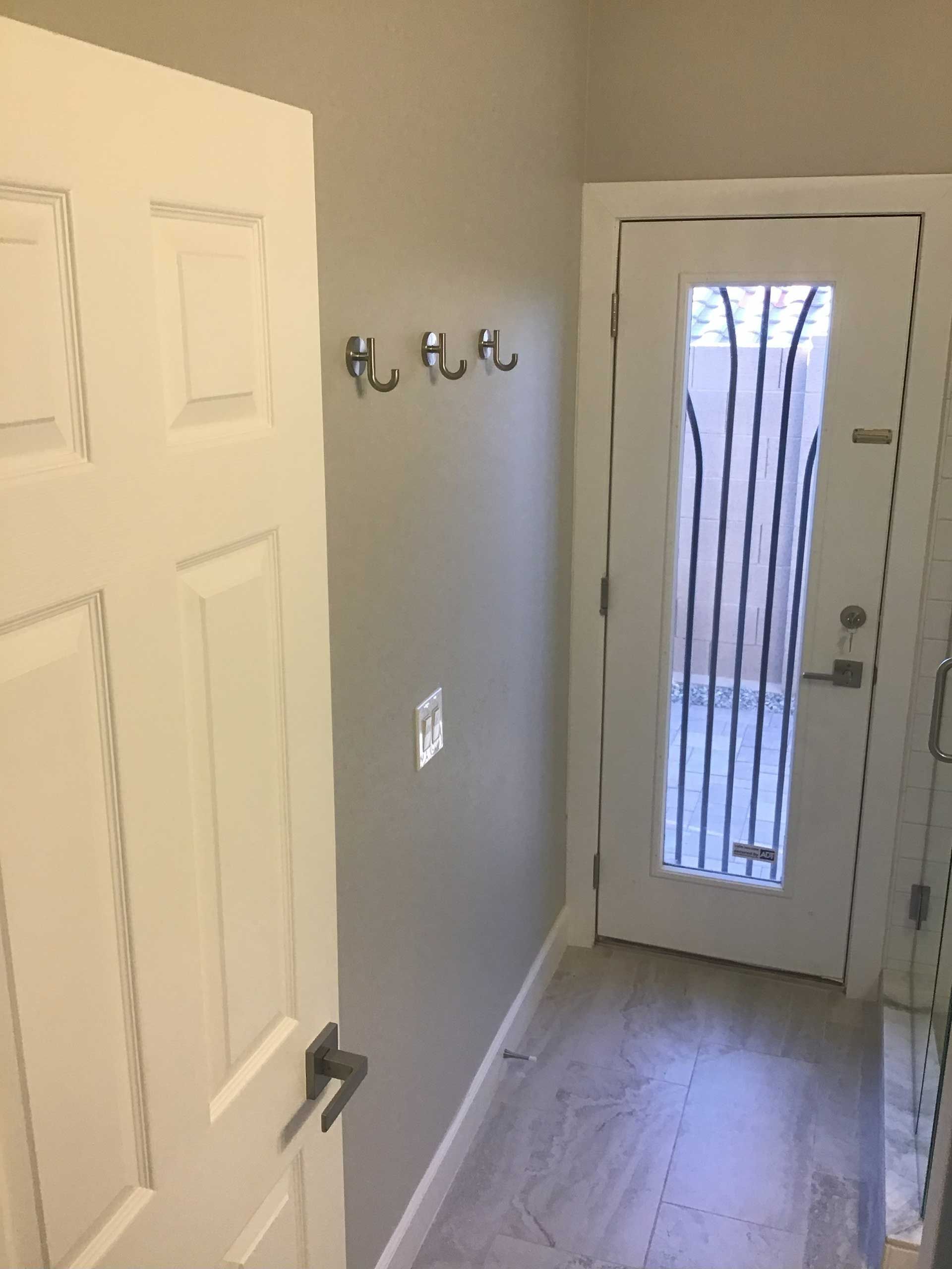 a narrow hallway with a closed white door on the left, three hooks on the gray wall, and a glass door to a patio on the right