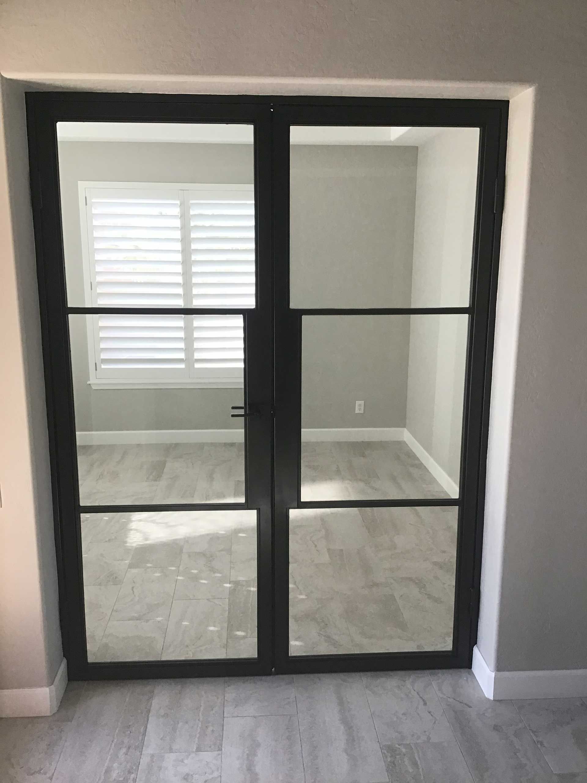black-framed glass French doors open to a room with gray walls, white shutters, and light flooring

