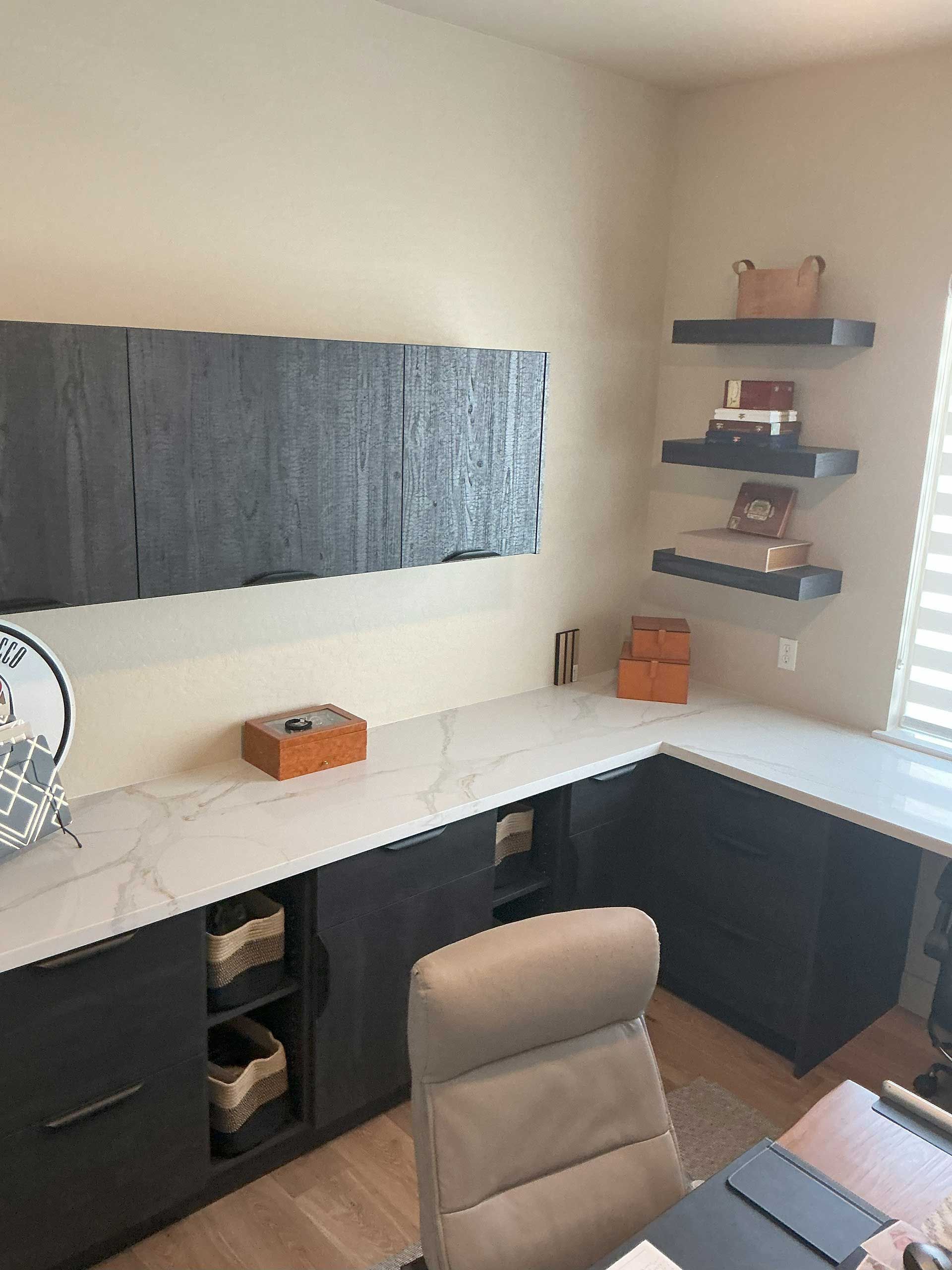 a modern home office with a white countertop, dark cabinets, and floating shelves holding designer boxes and books

