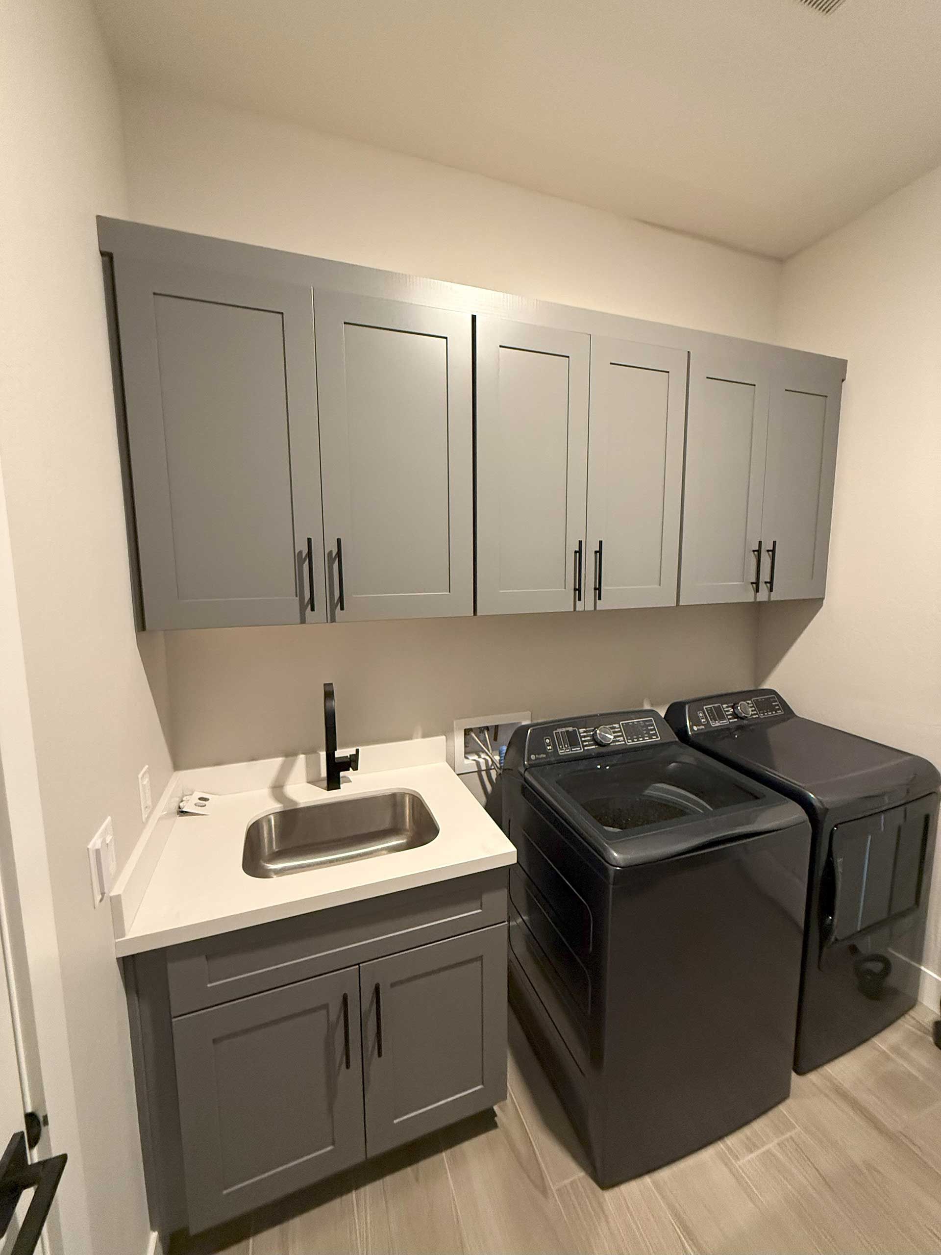 a laundry room with gray cabinets above a sink and dark-colored washer and dryer