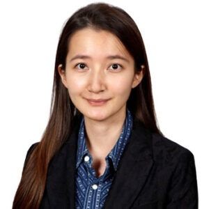 Woman with long brown hair wearing a dark blazer and blue patterned shirt, smiling.