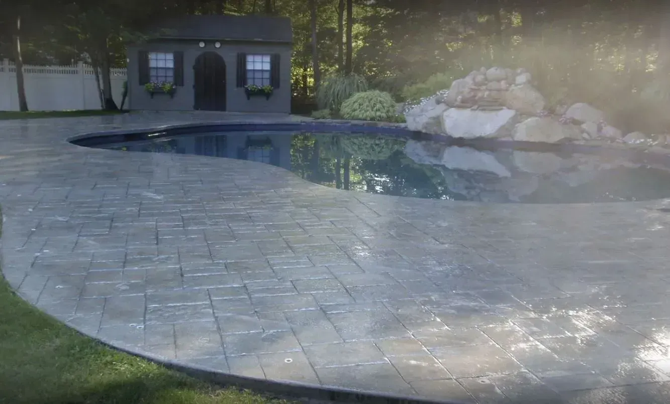 A sunlit backyard with a kidney-shaped pool, stamped concrete patio, a rock waterfall feature, and a small garden shed.