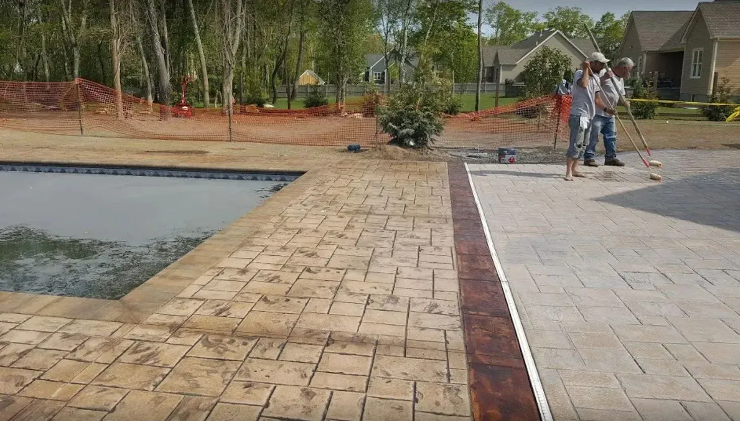Two workers spreading sand on a new patio next to a backyard swimming pool.