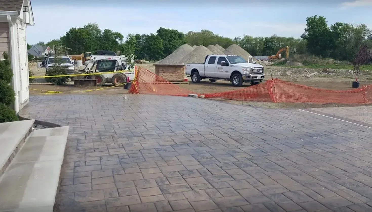 A paved patio overlooks a construction site with a truck, skid steer, and orange safety fencing under a cloudy sky.