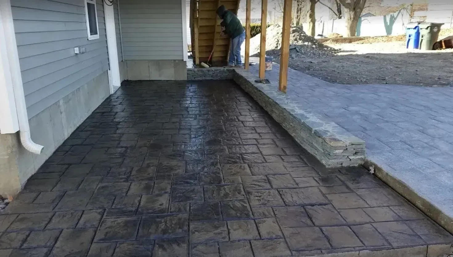 A stamped concrete patio in shades of brown borders a grey stone retaining wall under the edge of a house.
