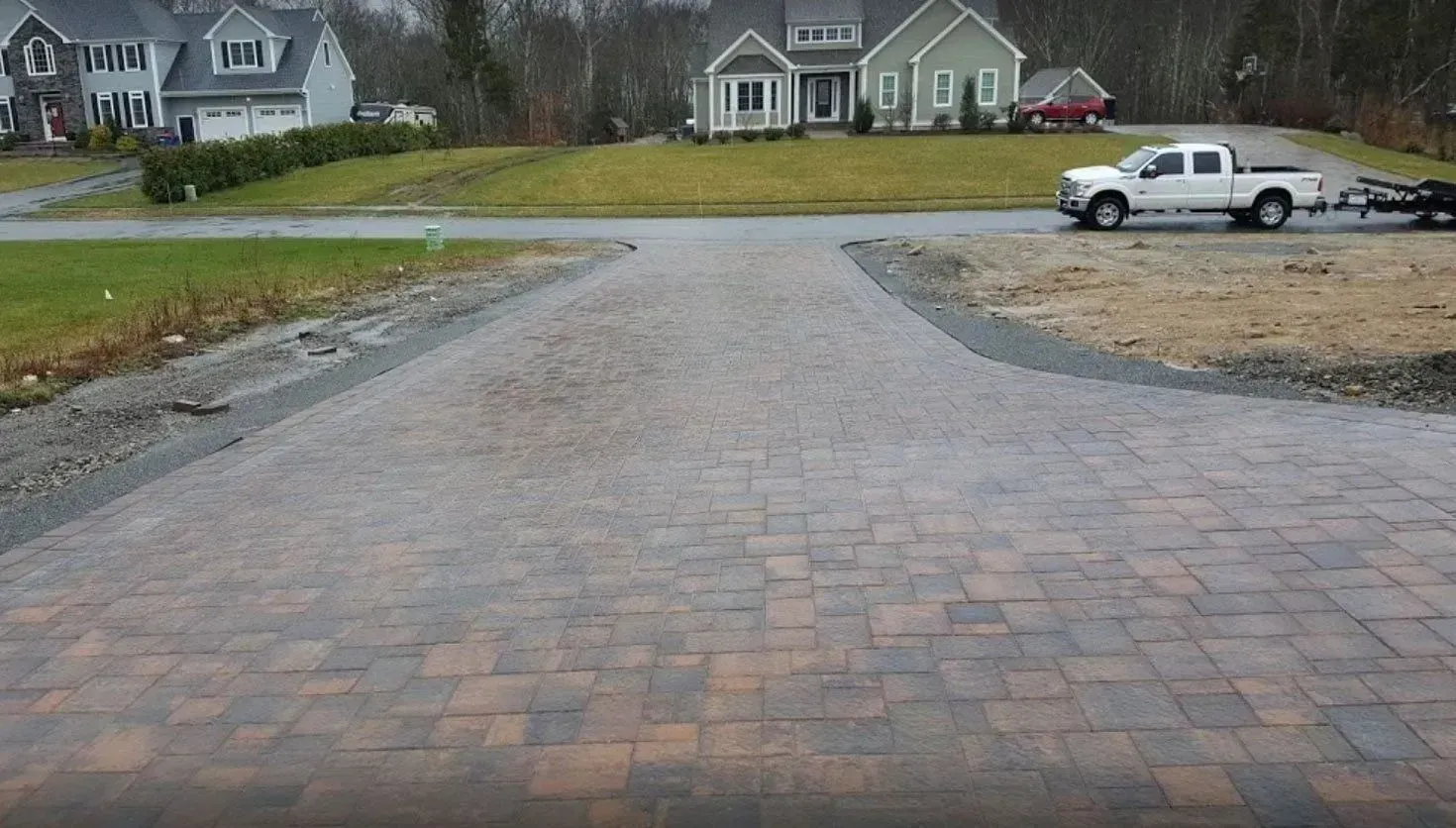 A newly paved stone driveway leads toward a quiet suburban street with two houses and a white pickup truck in the distance.