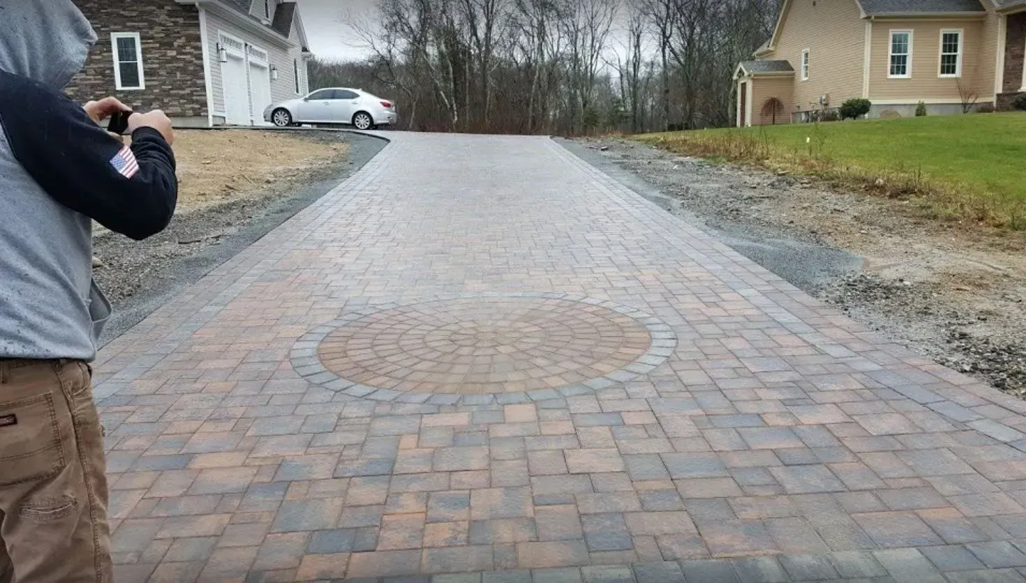 A person photographs a new paver driveway with a central decorative circle, leading toward two houses in a residential area.