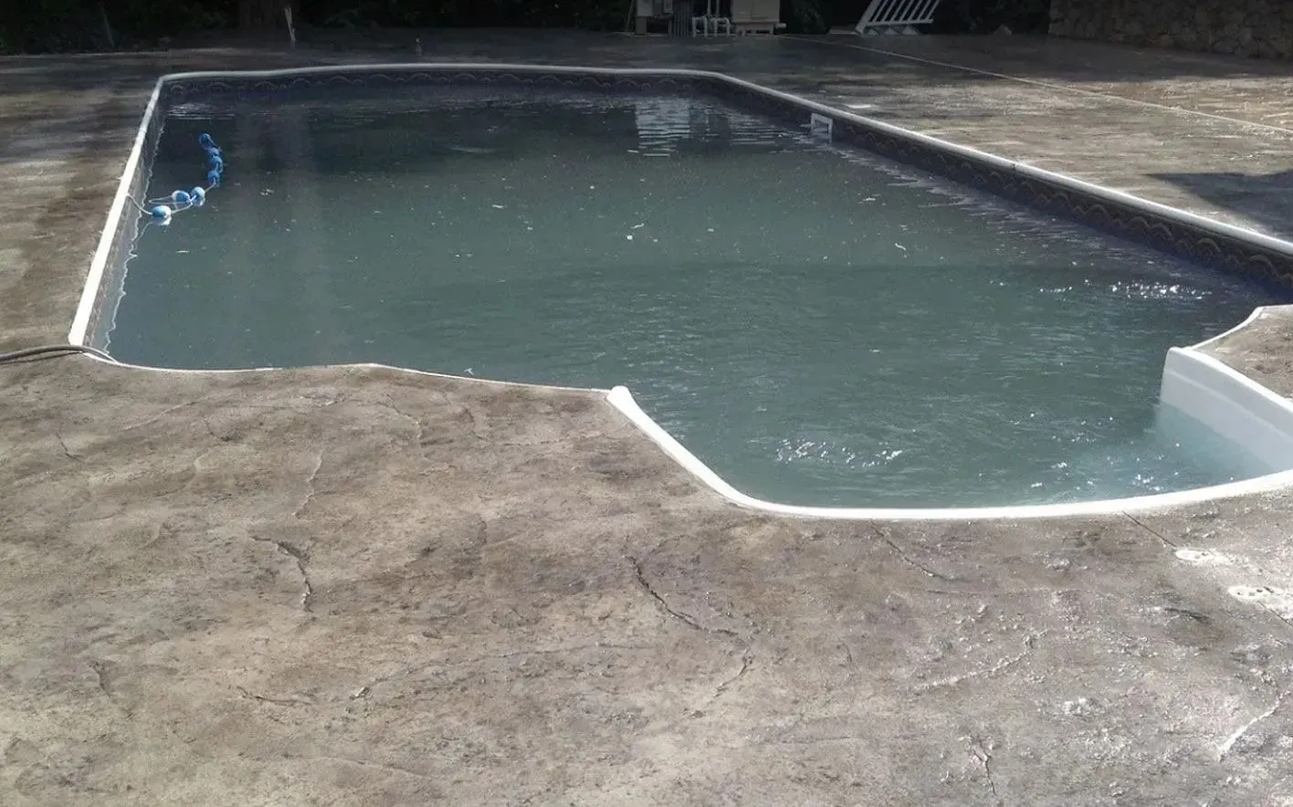 An outdoor swimming pool with gray-toned water, surrounded by textured, weathered concrete decking.