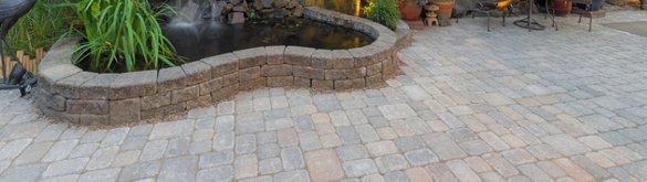 A small, curved, stone-walled garden pond sits beside a patio paved with light, multi-colored rectangular pavers.