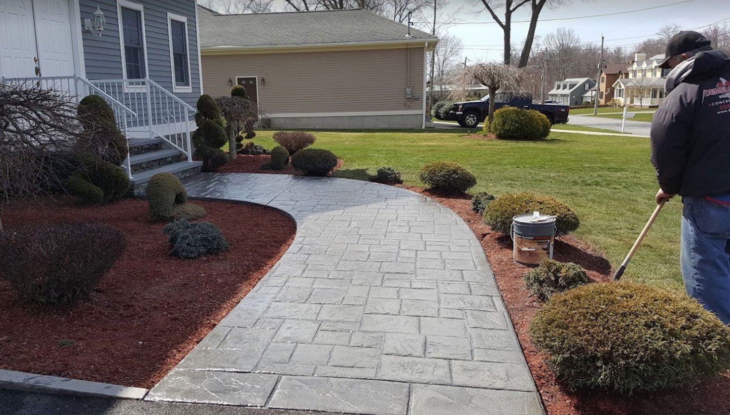 A worker cleans the edges of a curved, gray stamped concrete walkway beside a house with red mulch landscaping.