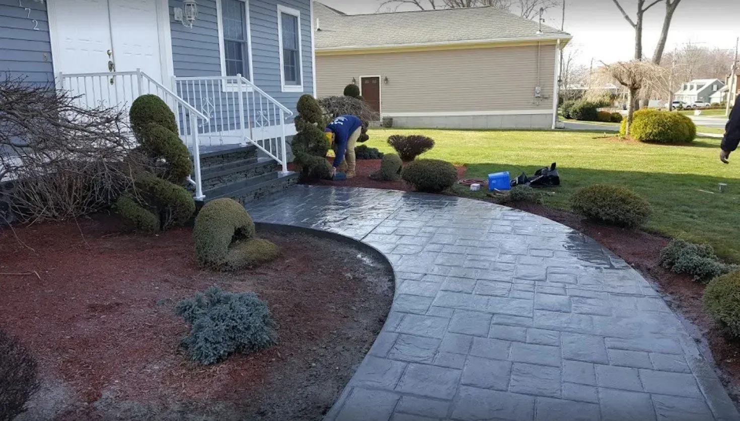A person works on landscaping in front of a blue-sided house with a newly installed gray stone walkway.
