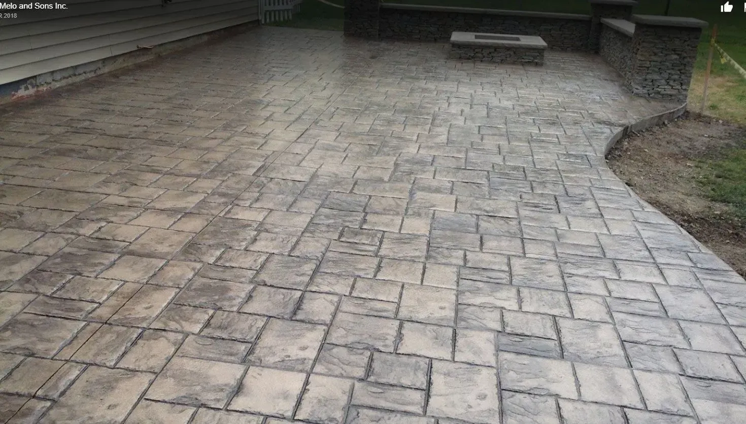 A gray, patterned paver patio with a stone wall and built-in fire pit, adjacent to a house exterior.