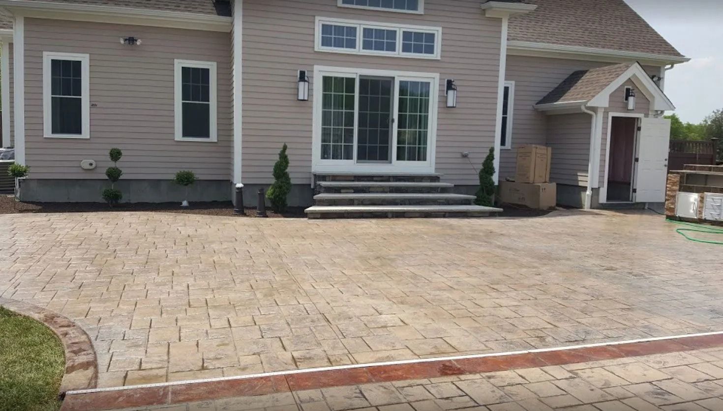 A paved patio leading to the sliding glass doors and side entrance of a light-colored, two-story suburban home.