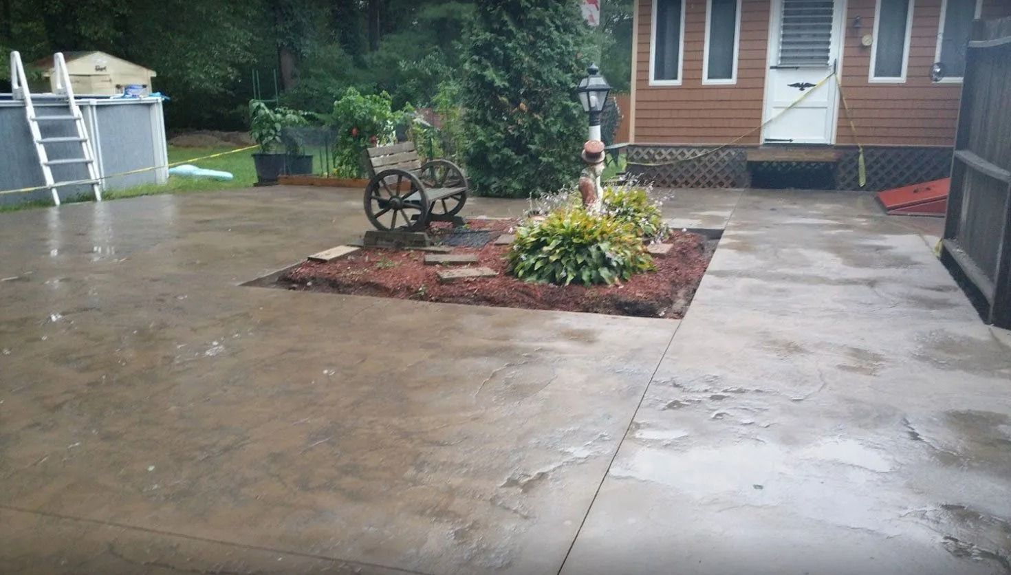 A concrete patio features a small central garden bed with a wooden wagon-wheel bench, shrubs, and a lamppost.