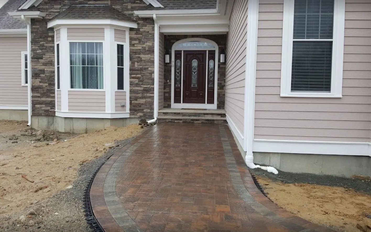 A paver walkway leads to the front entrance of a house with light-colored siding, a stone facade, and a bay window.