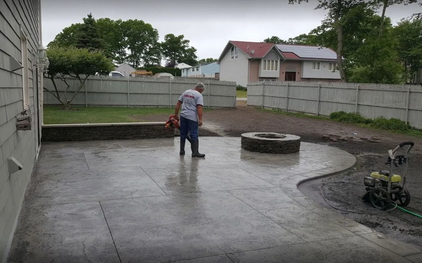A person in a gray shirt stands on a finished gray stamped concrete patio with a circular fire pit in a backyard.