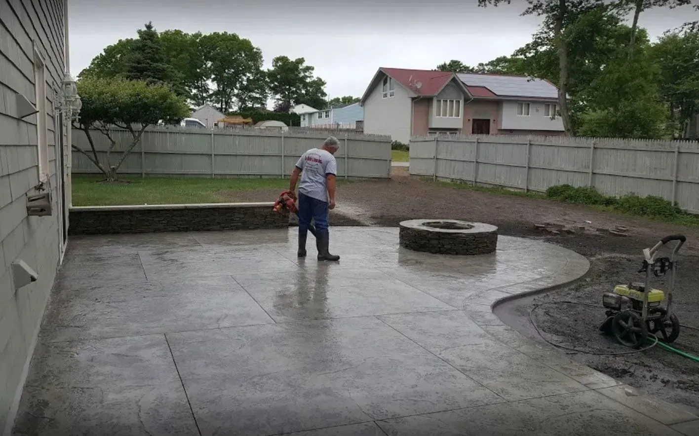 A person uses a leaf blower on a newly installed stamped concrete patio with a stone fire pit in a residential backyard.