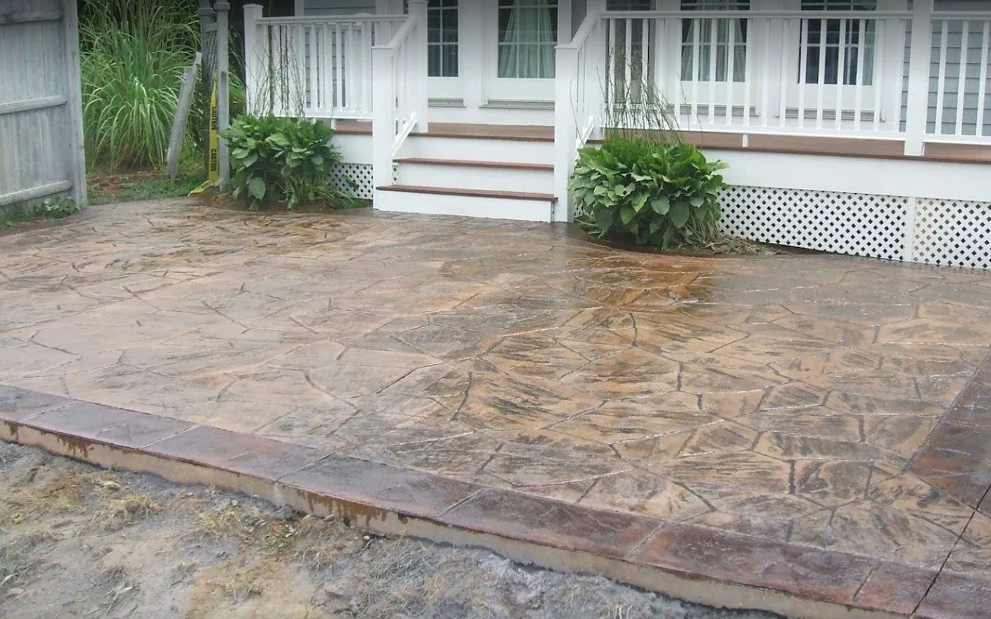 A stamped, tan-colored concrete patio with a dark border, located in front of a white house with a porch and steps.