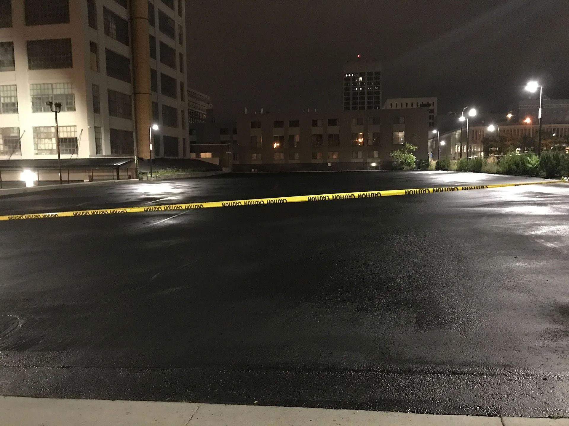 A night view of a dark, empty parking lot cordoned off by yellow caution tape, with city buildings illuminated in the back.