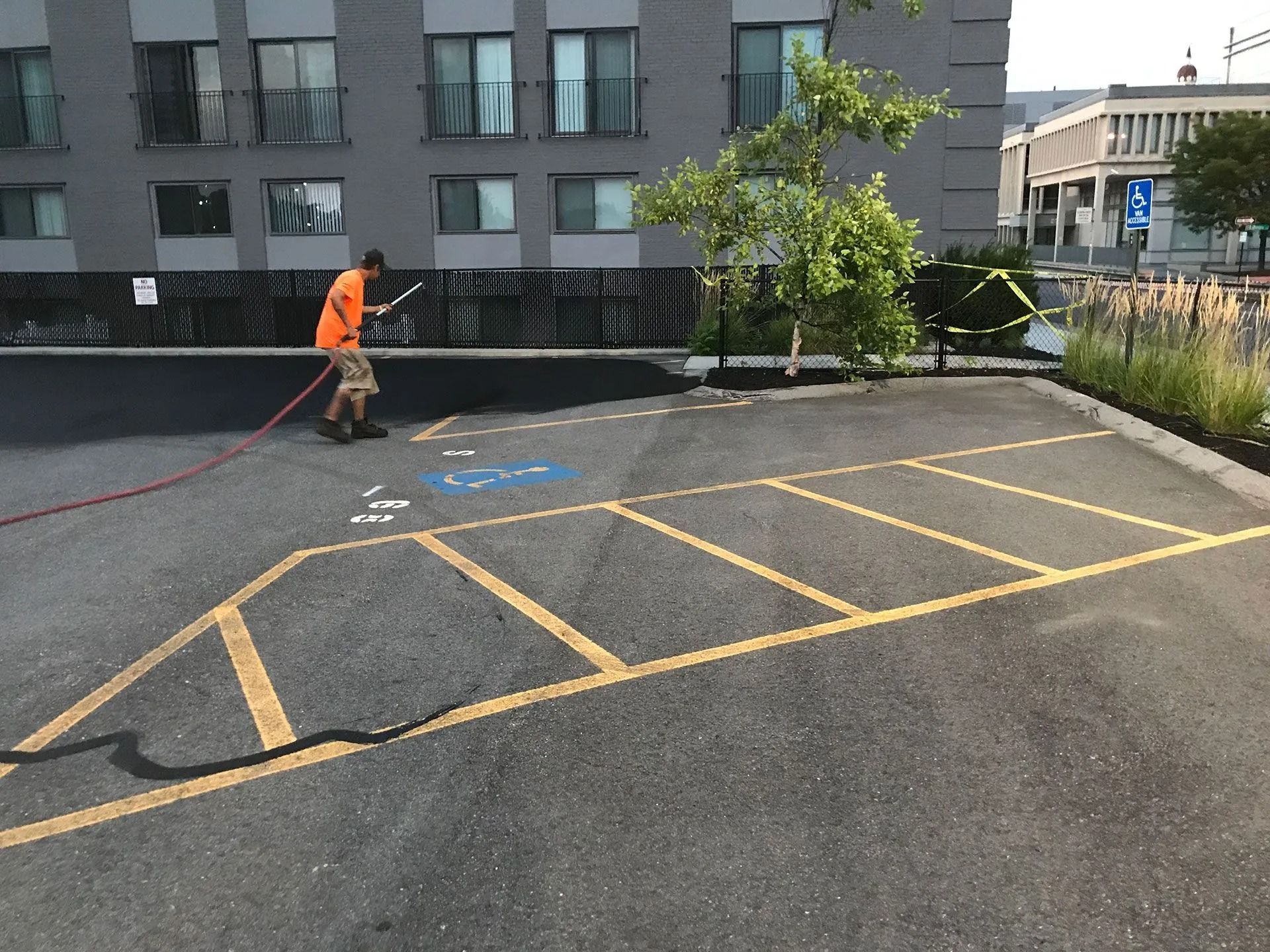 A person in an orange shirt uses a spray hose to sealcoat a parking lot with yellow-striped spaces and a blue icon.