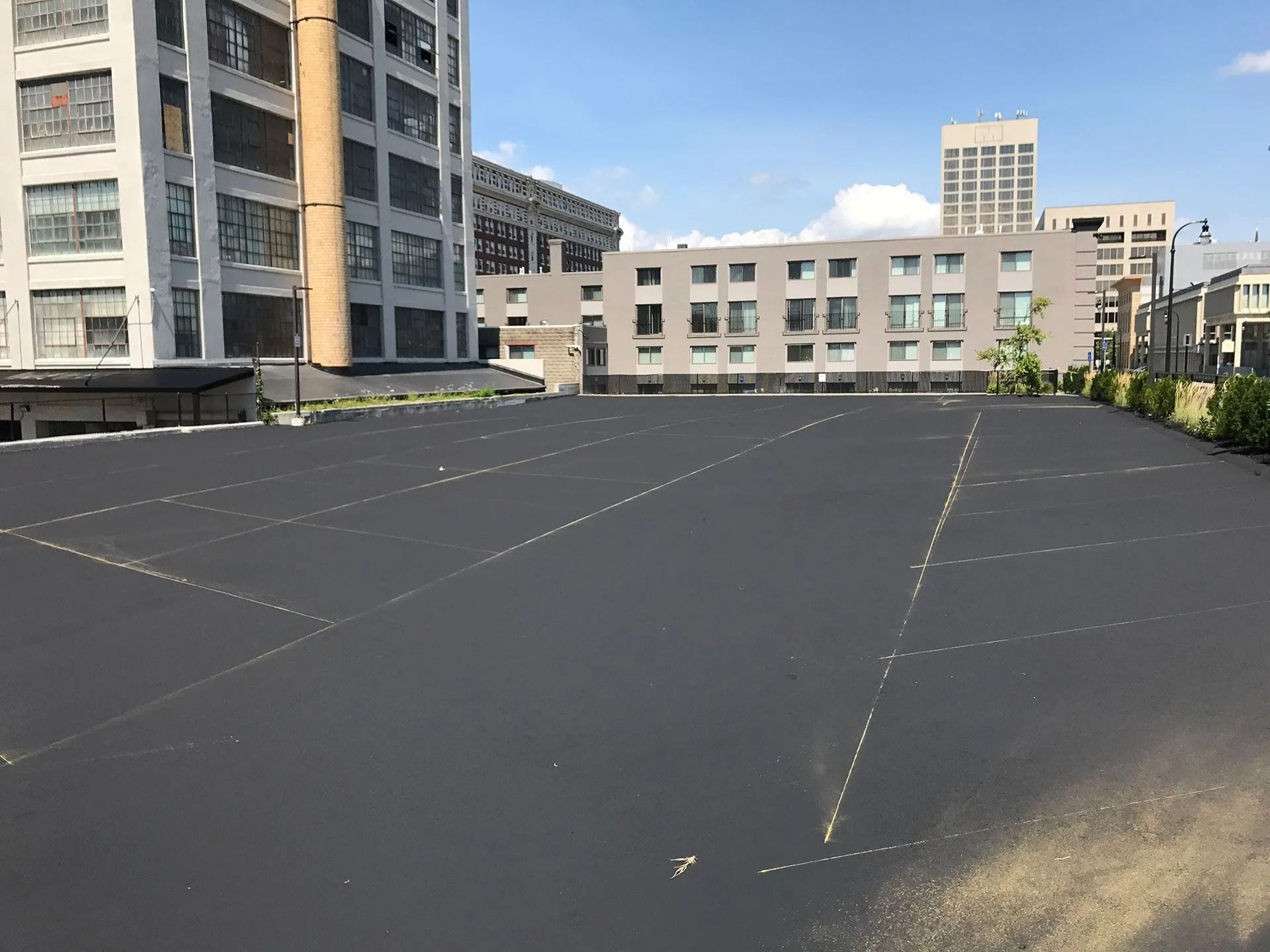 A newly paved, empty asphalt parking lot with white painted stall lines, flanked by multi-story city buildings.