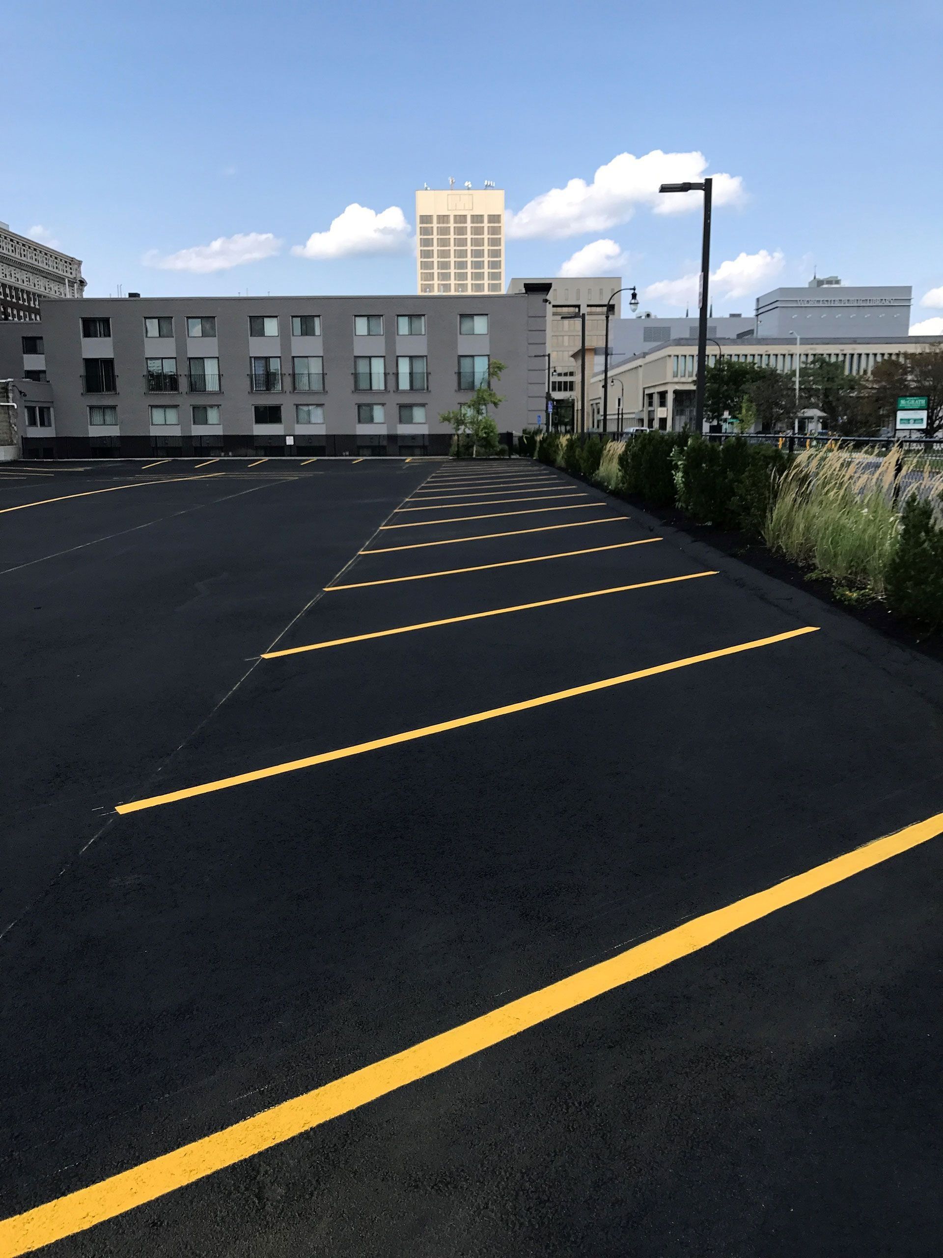 A freshly paved parking lot with yellow painted lines and a row of green bushes against a backdrop of city buildings.