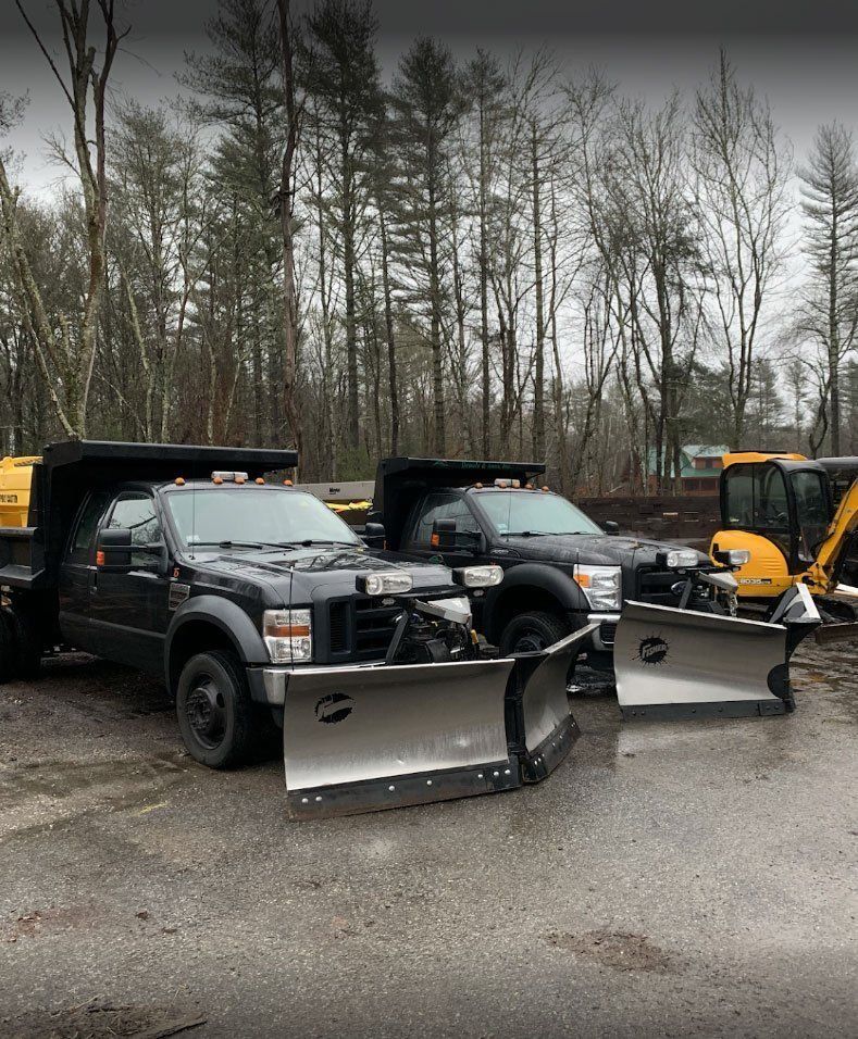 Two dark-colored dump trucks with snow plows attached, parked on a gravel lot in front of a forest.