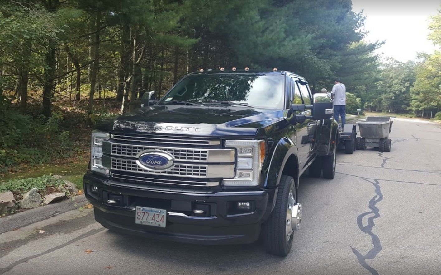 A black Ford Super Duty truck parked on an asphalt road next to a wooded area, with a person standing near a small trailer.