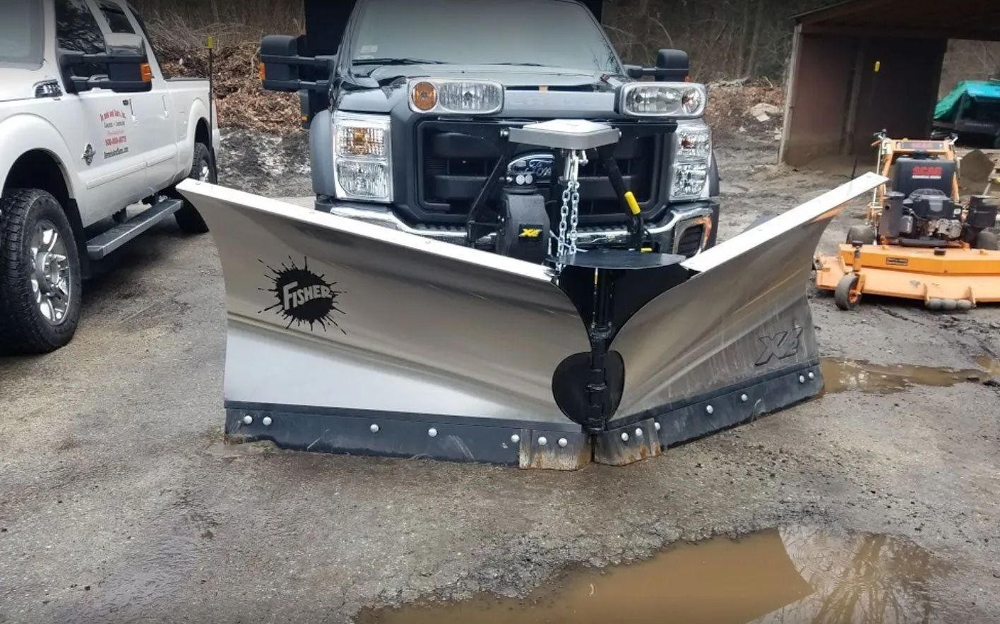 A gray Ford truck with a large Fisher V-plow mounted on the front, parked on a gravel lot near a lawn mower.