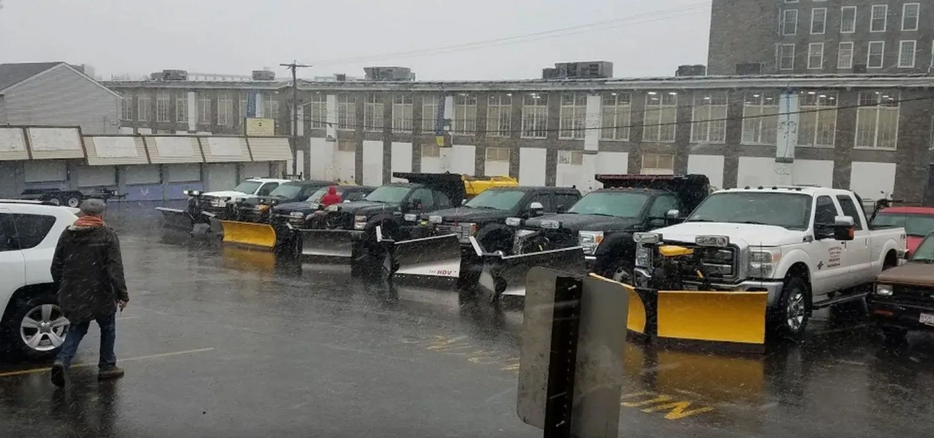 A row of pickup trucks equipped with yellow snowplows parked in a wet, paved lot in front of a brick building.
